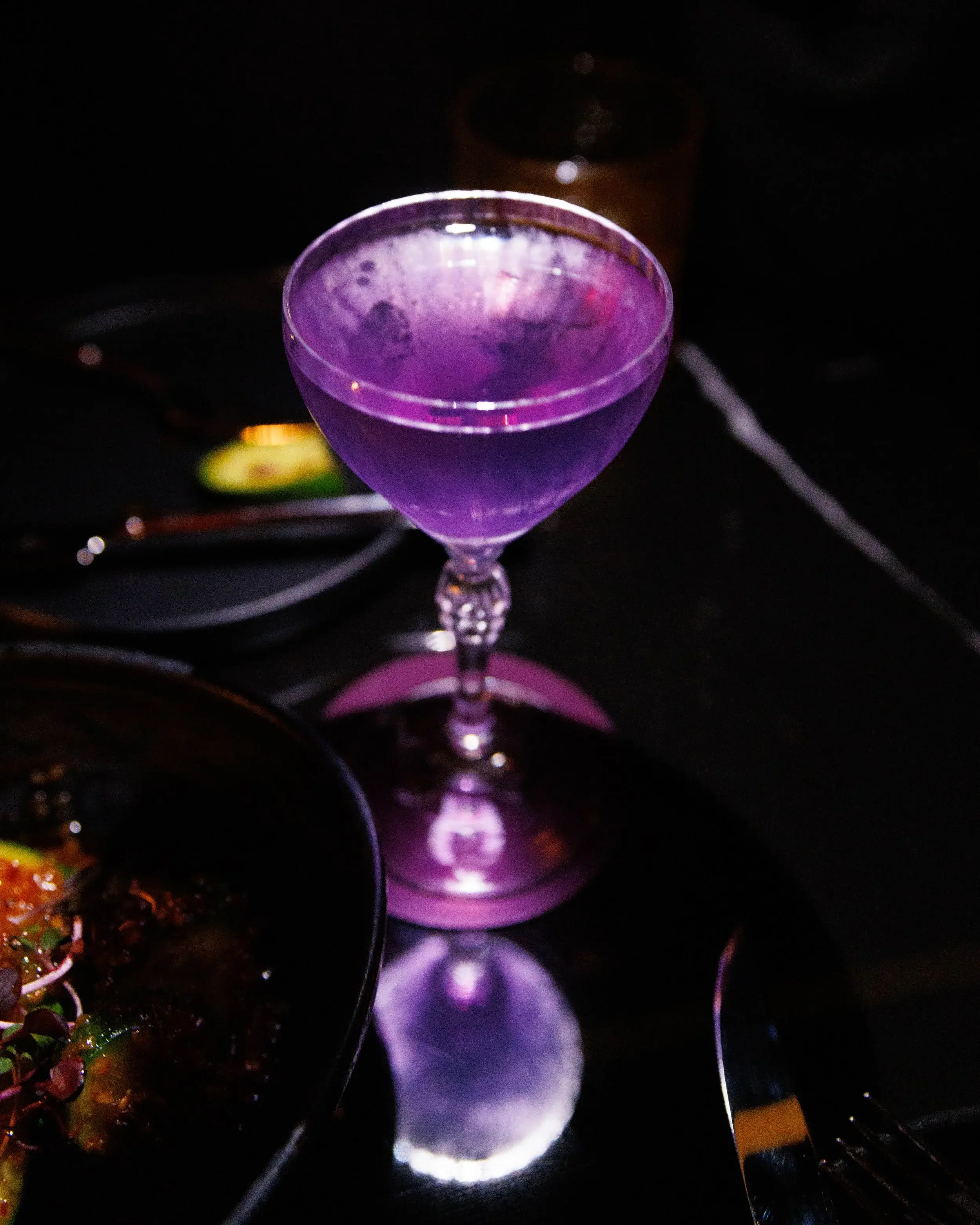 Purple cocktail in a stemmed coupe reflecting light on a black tabletop