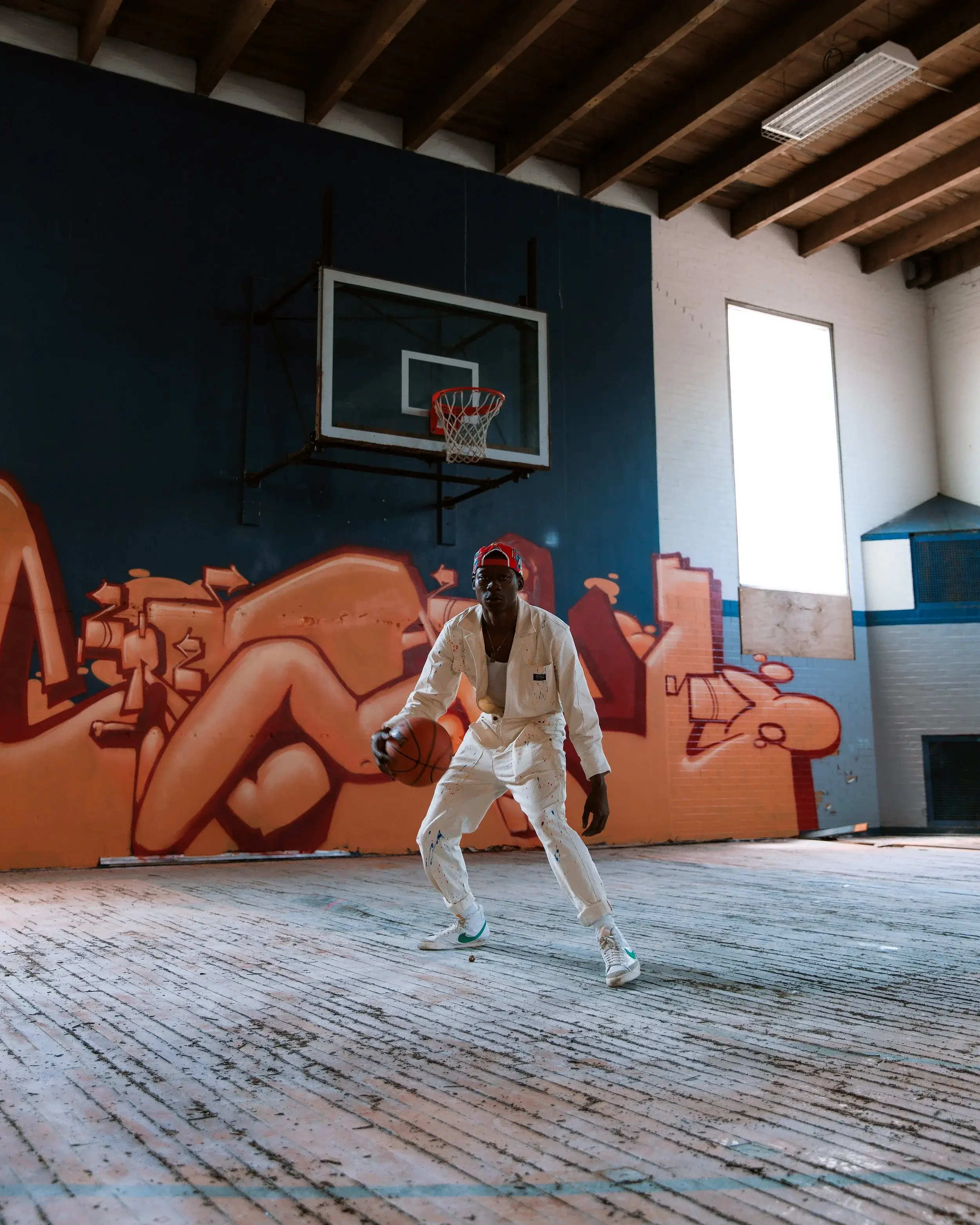Model in a white workwear jumpsuit dribbling a basketball in a graffiti-covered gym