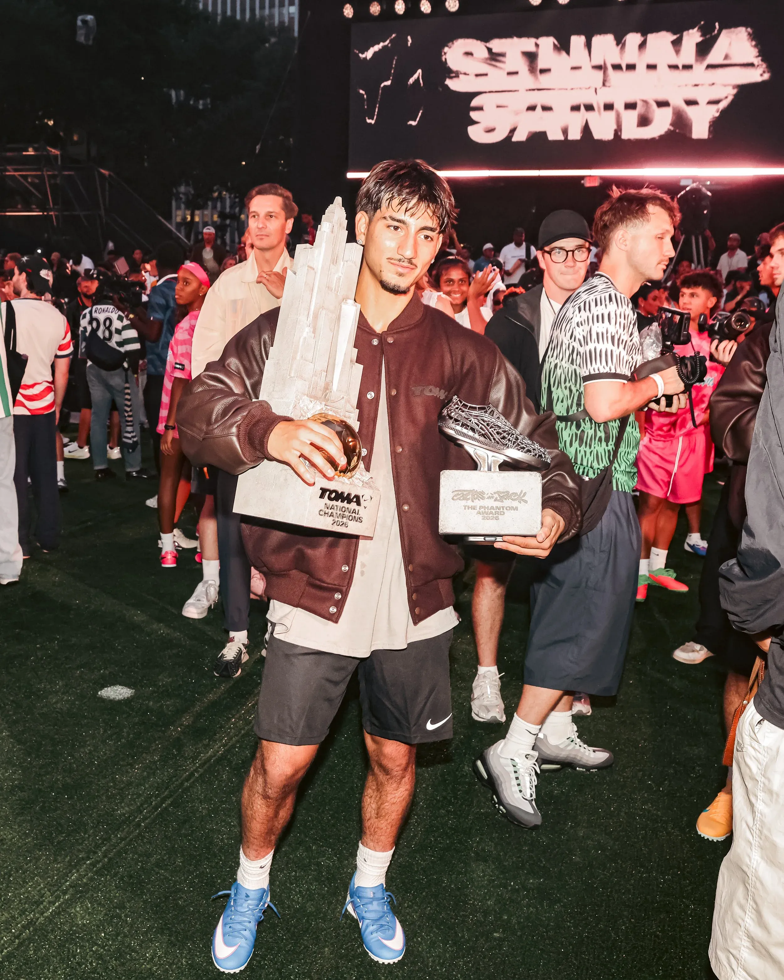 Competitor holding two trophies on a turf field at a crowded evening event