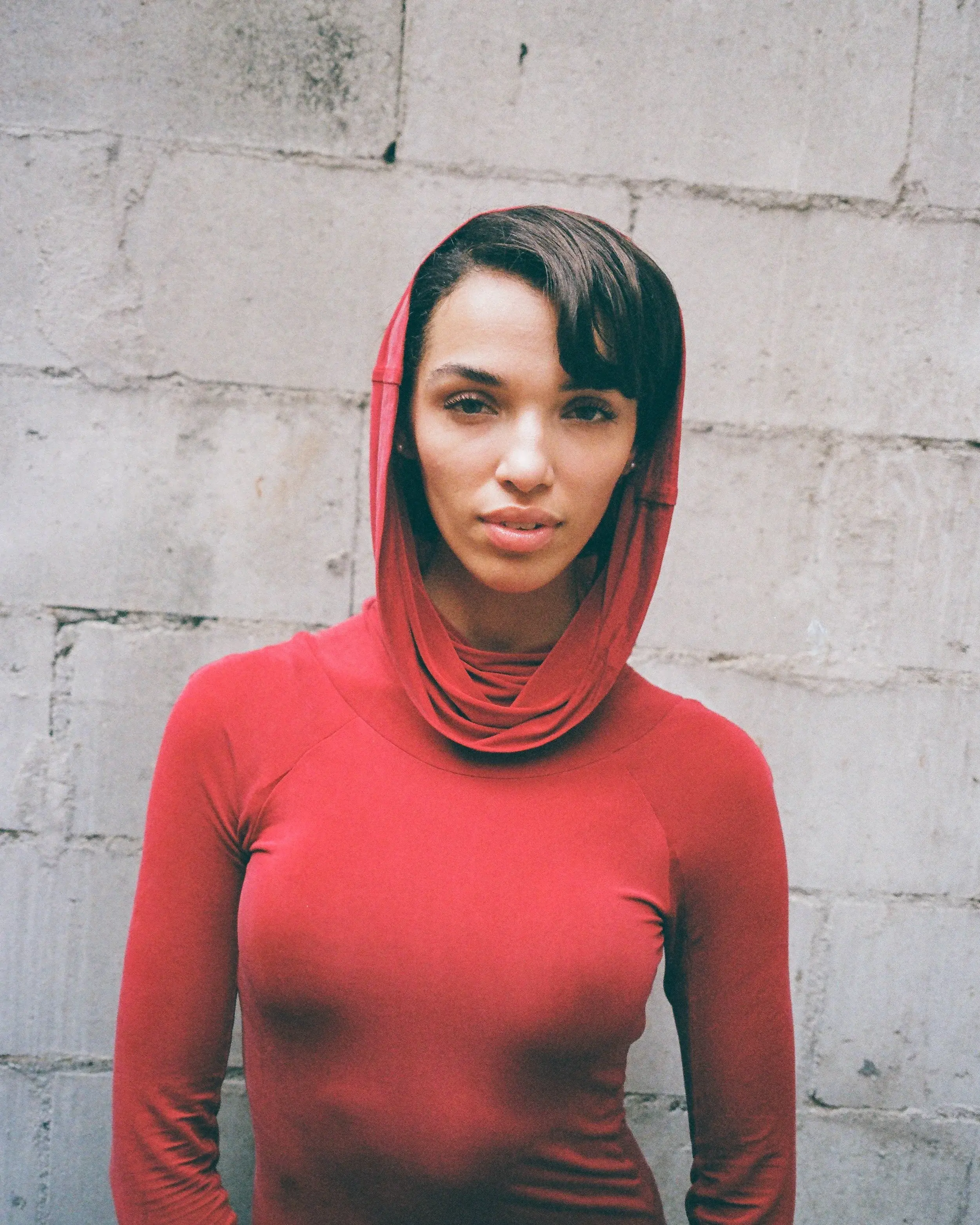 Woman in a red hooded top gazing at the camera against a gray block wall