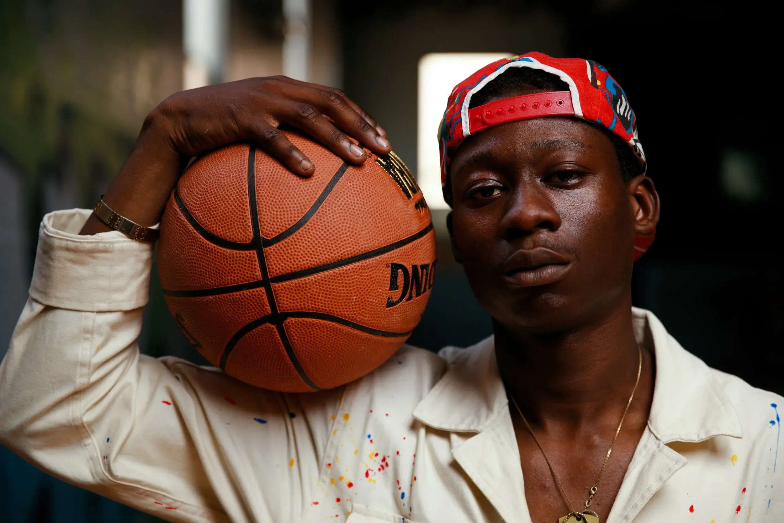Campaign portrait of a model in a red cap and paint-splattered shirt holding a basketball