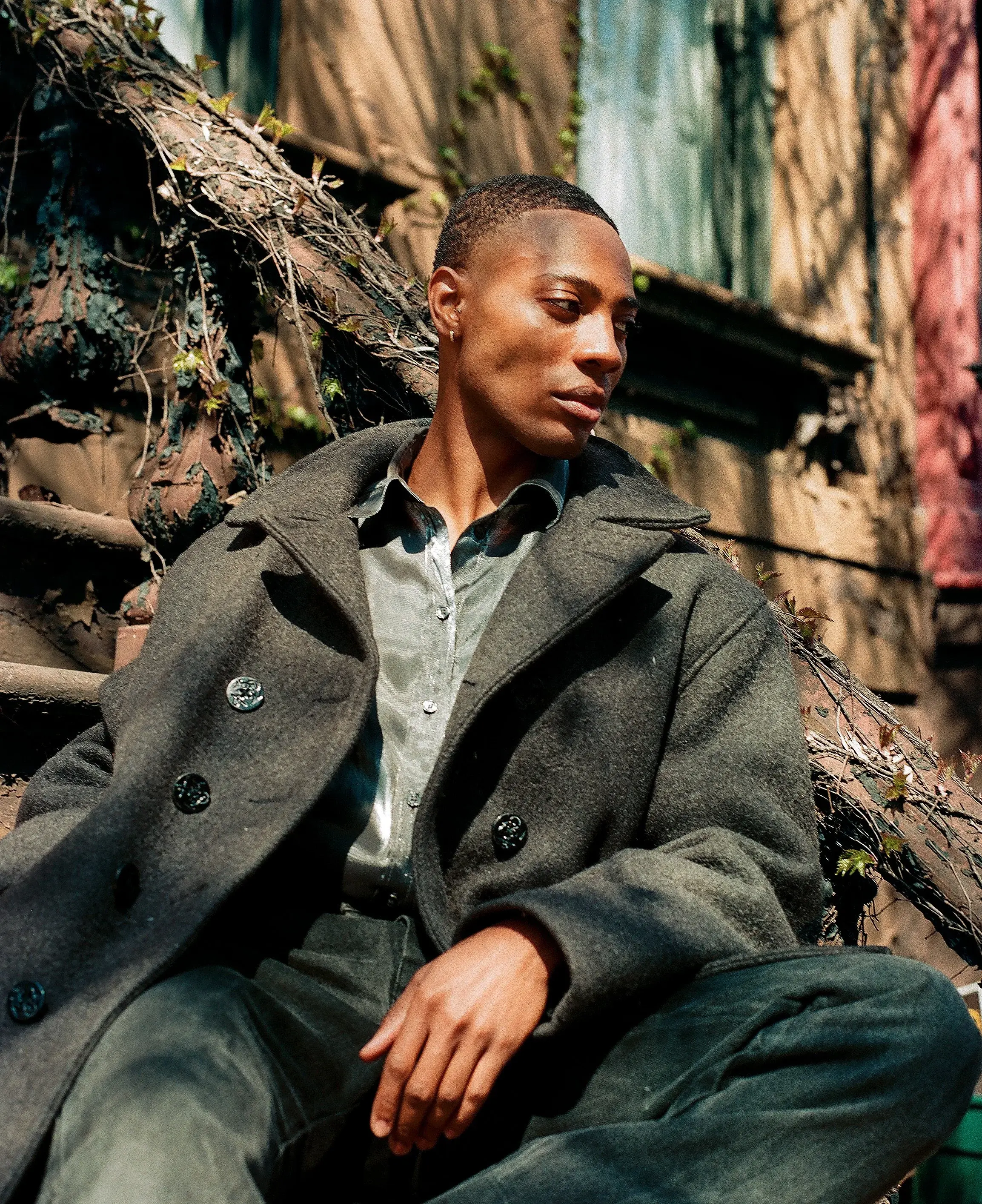 Man in a dark wool overcoat sitting among tangled vines, looking away in warm sunlight