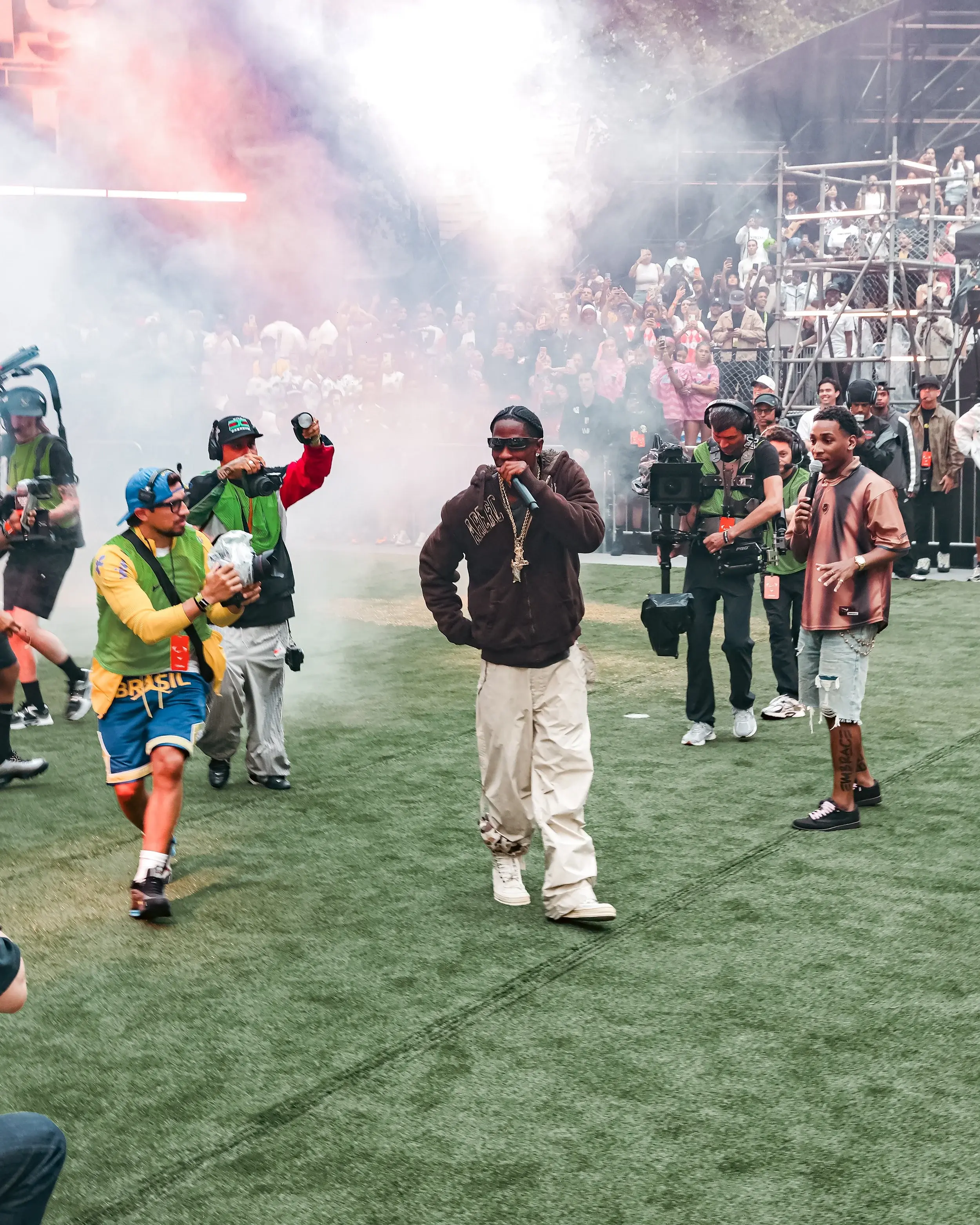 Performers with microphones walking through smoke on a turf field before a packed crowd