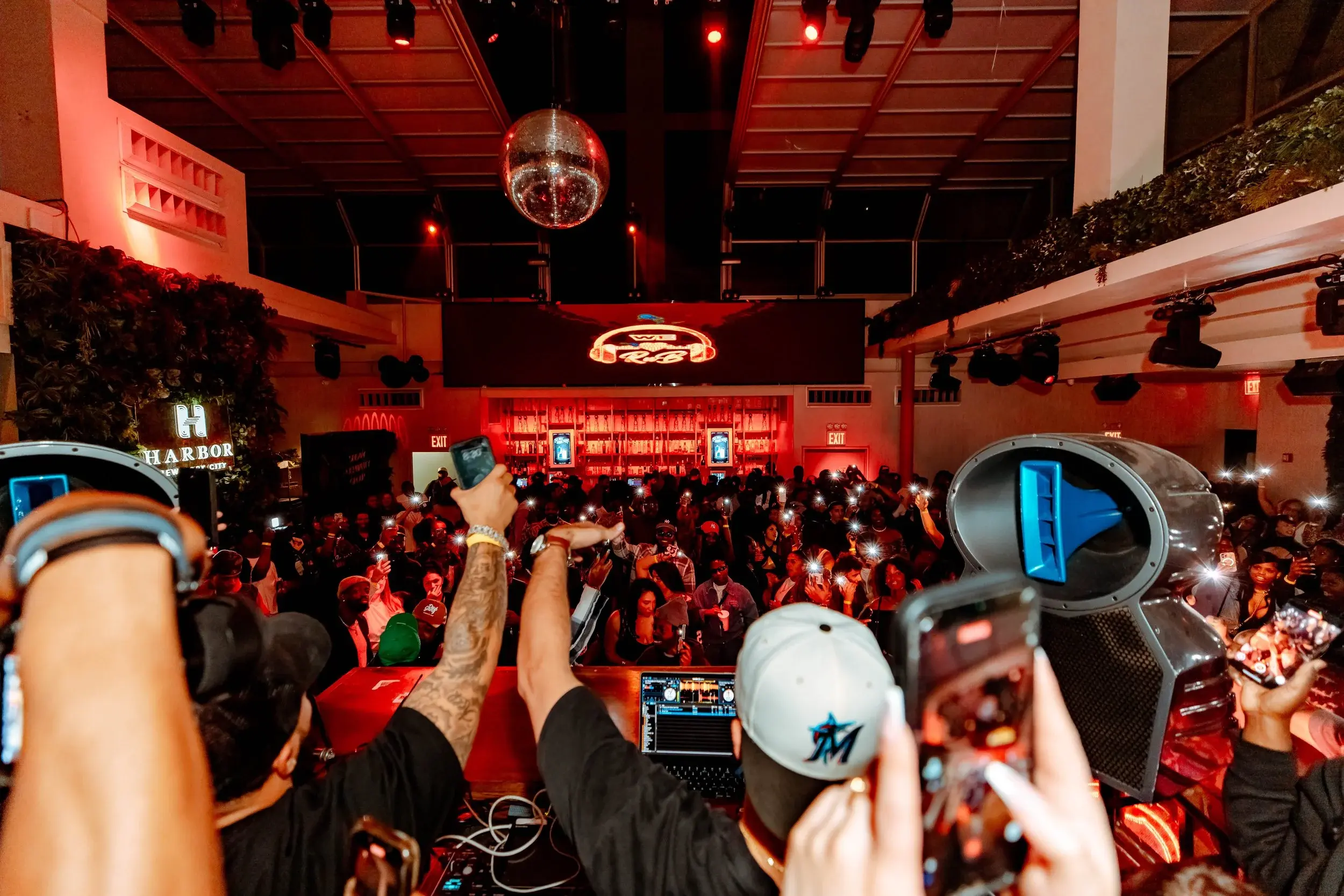 Wide view of a red-lit nightlife crowd raising phones beneath a disco ball