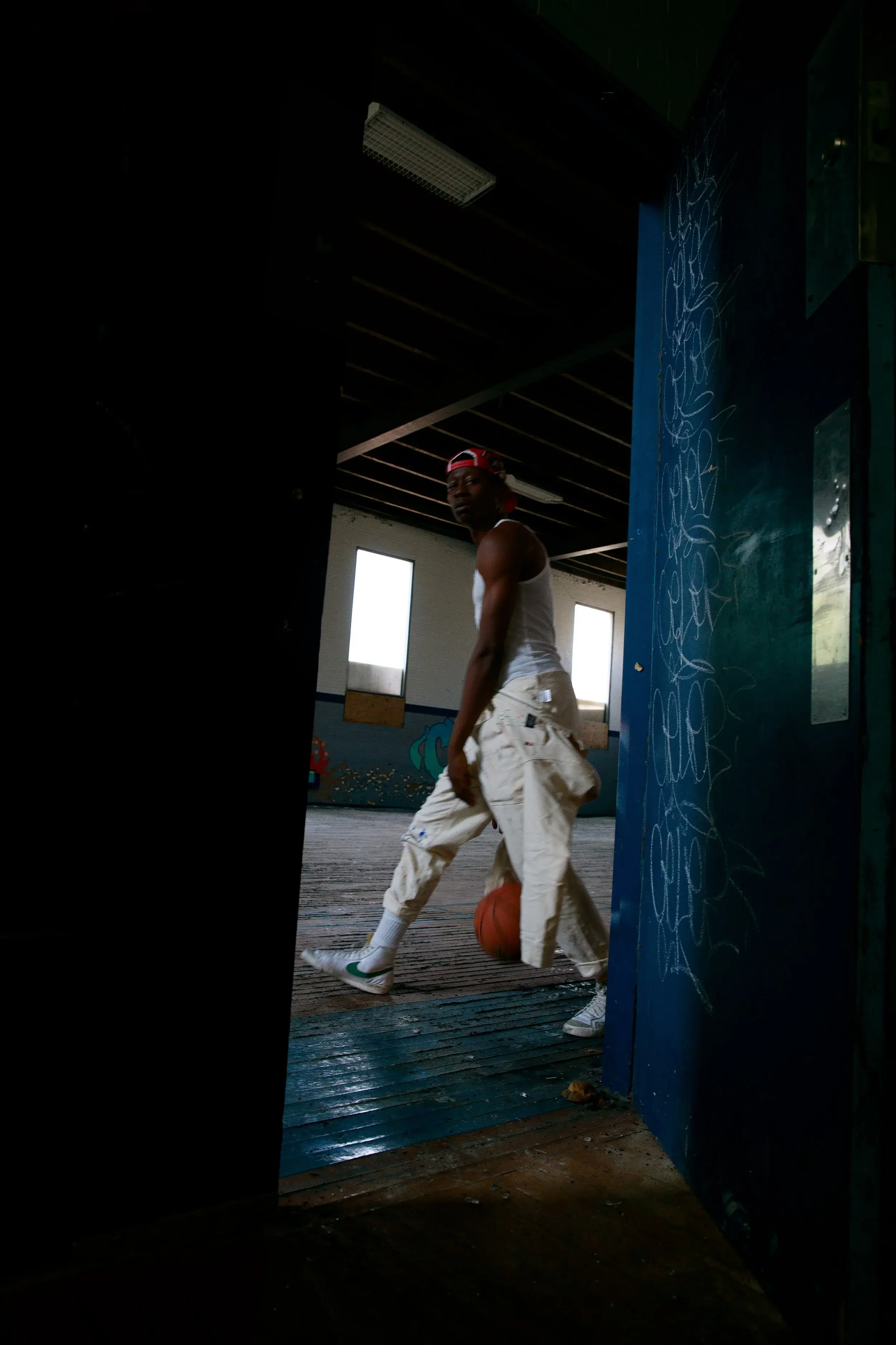 Streetwear model dribbling a basketball across a dim gym, seen through a blue doorway