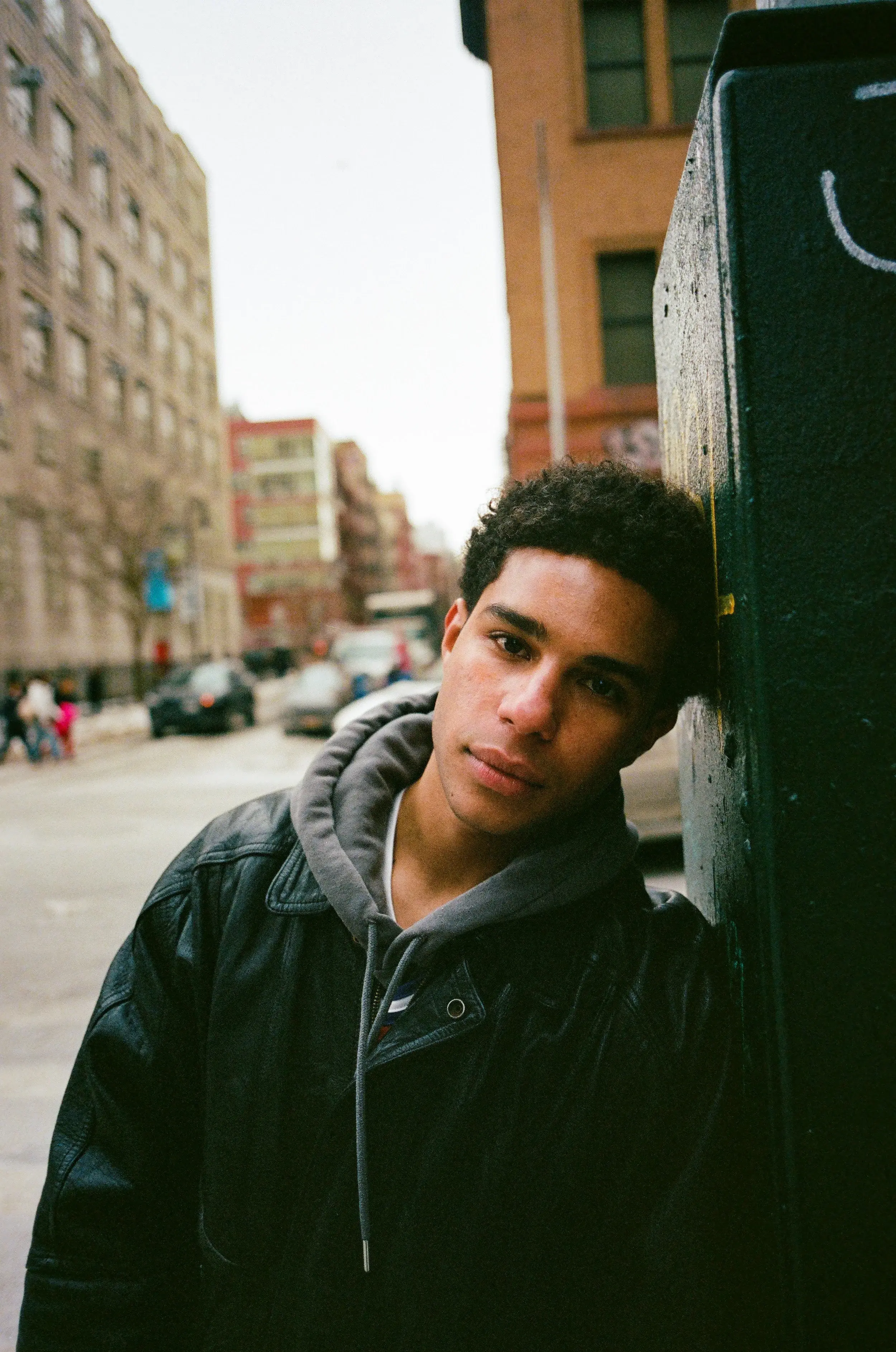 Young man in a leather jacket and gray hoodie leaning against a dark pillar