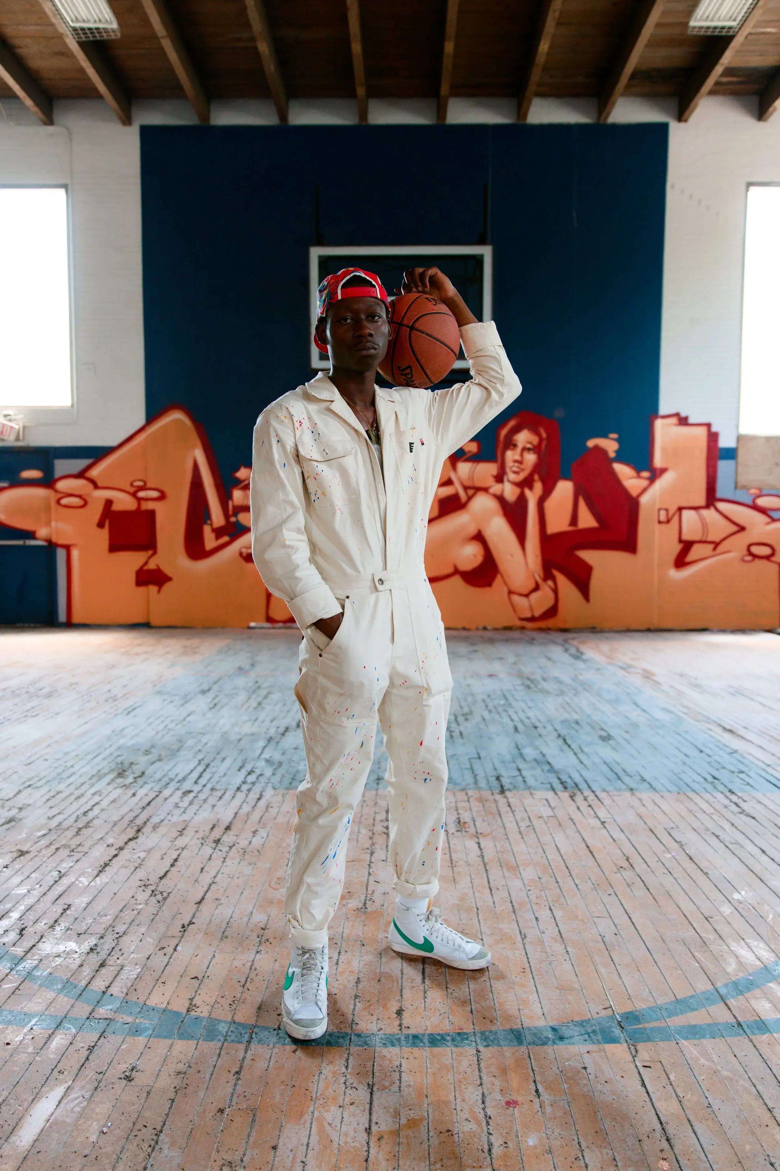 Model in a white coverall holding a basketball on one shoulder before a graffiti mural