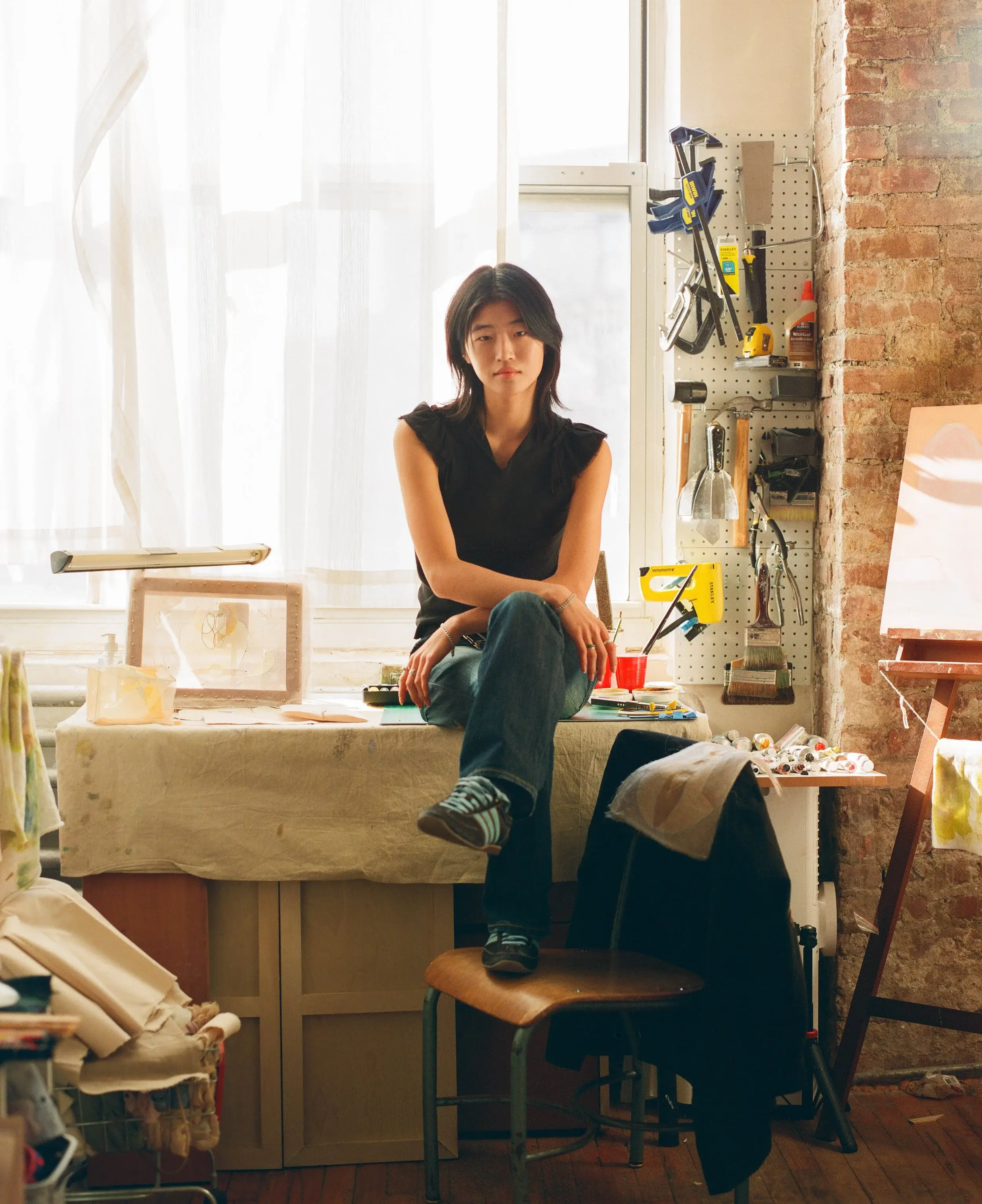 Relaxed portrait of a woman perched on a worktable surrounded by tools and sunlight