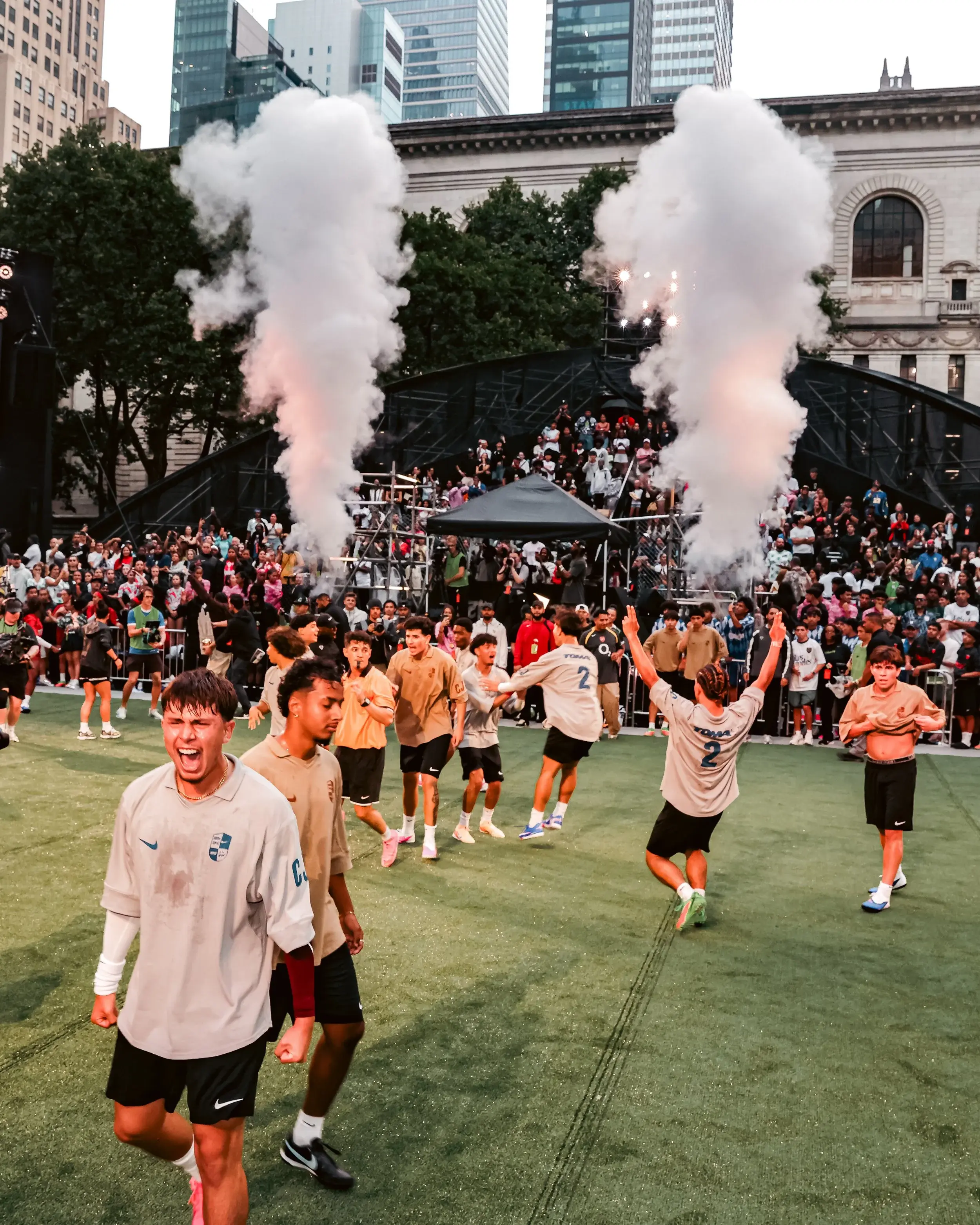 Players celebrating with raised arms as smoke plumes rise over an outdoor competition