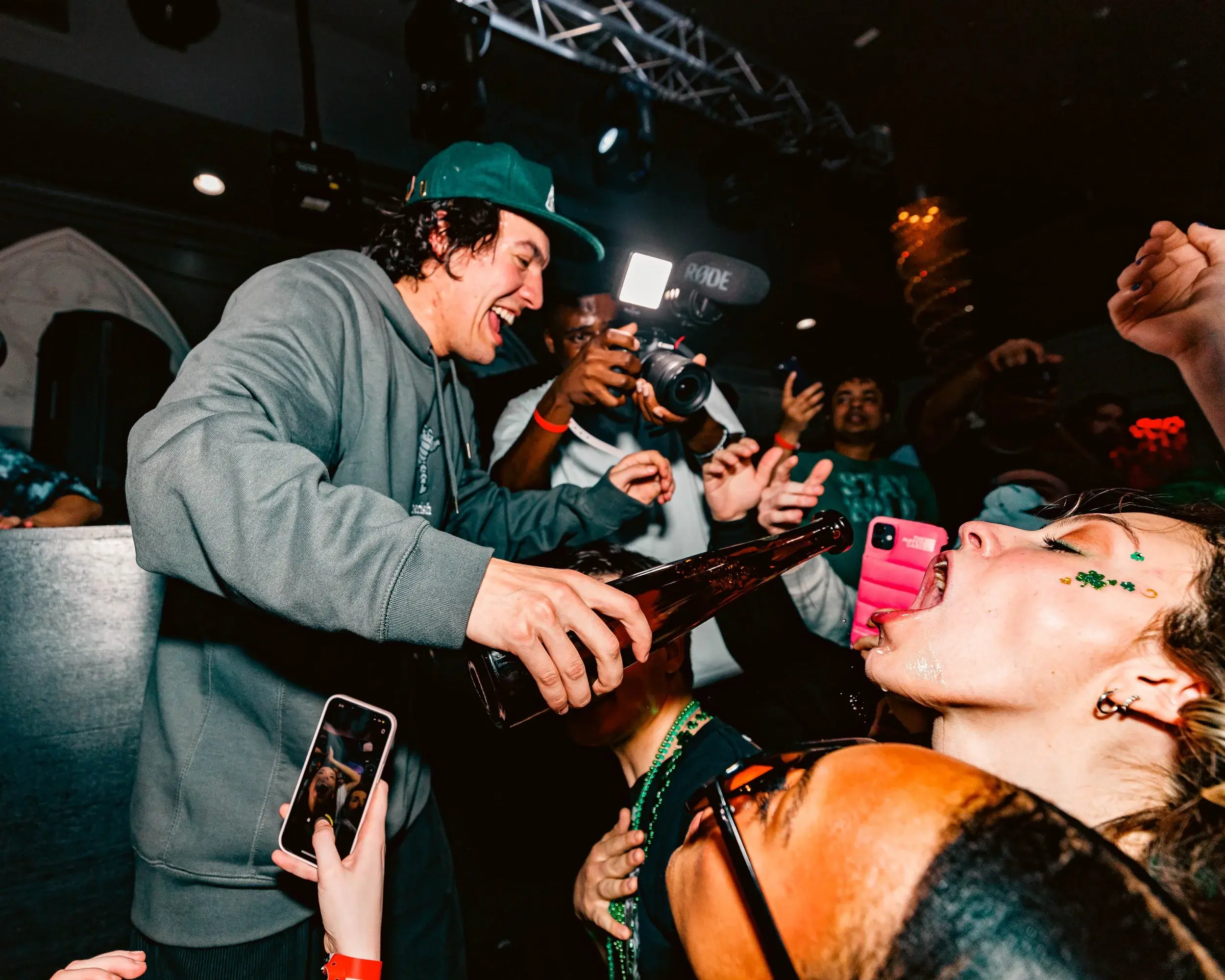Man pouring a drink into a friend's mouth as cameras flash around them