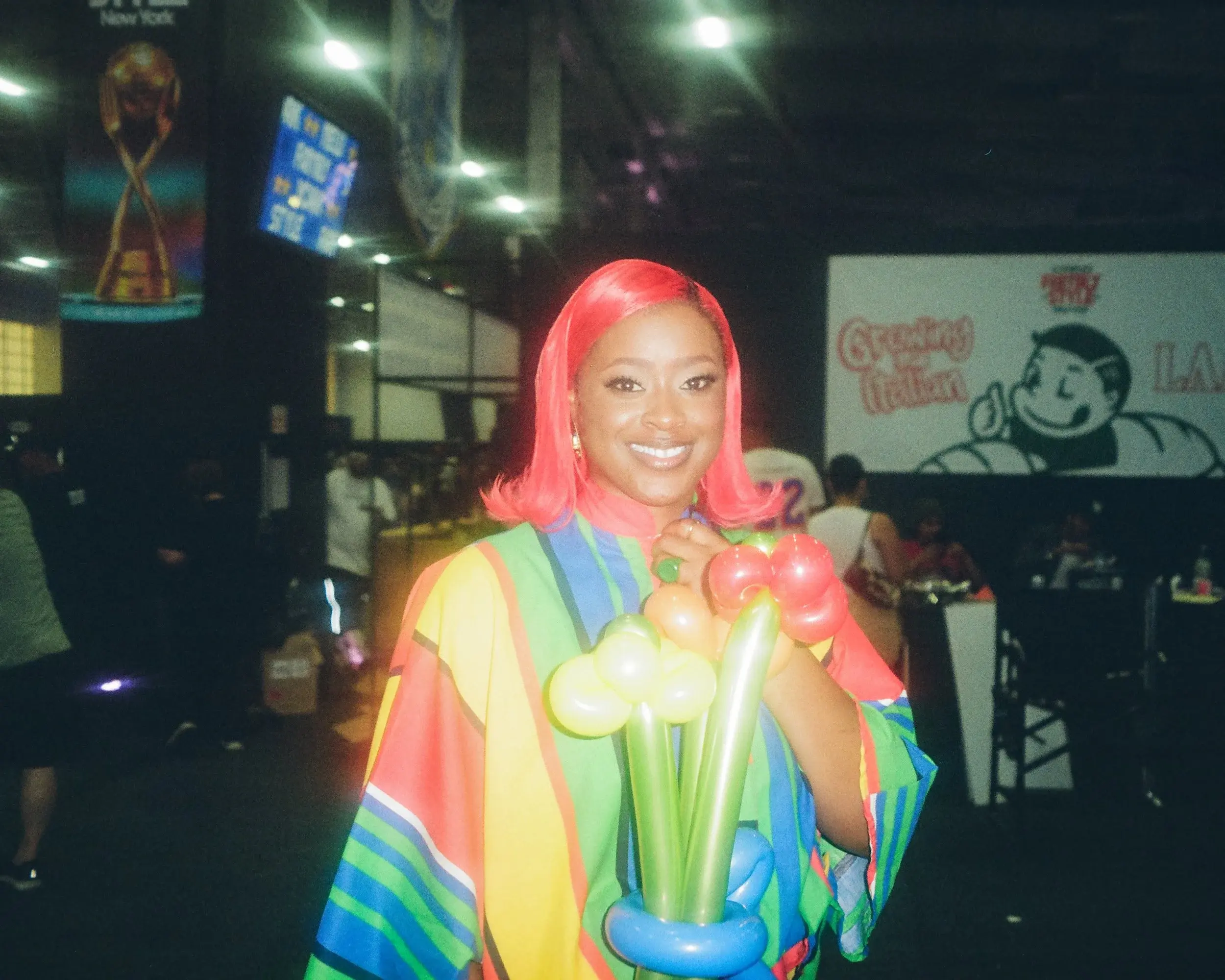 Smiling woman with bright pink hair holding balloon flowers in a busy indoor hall