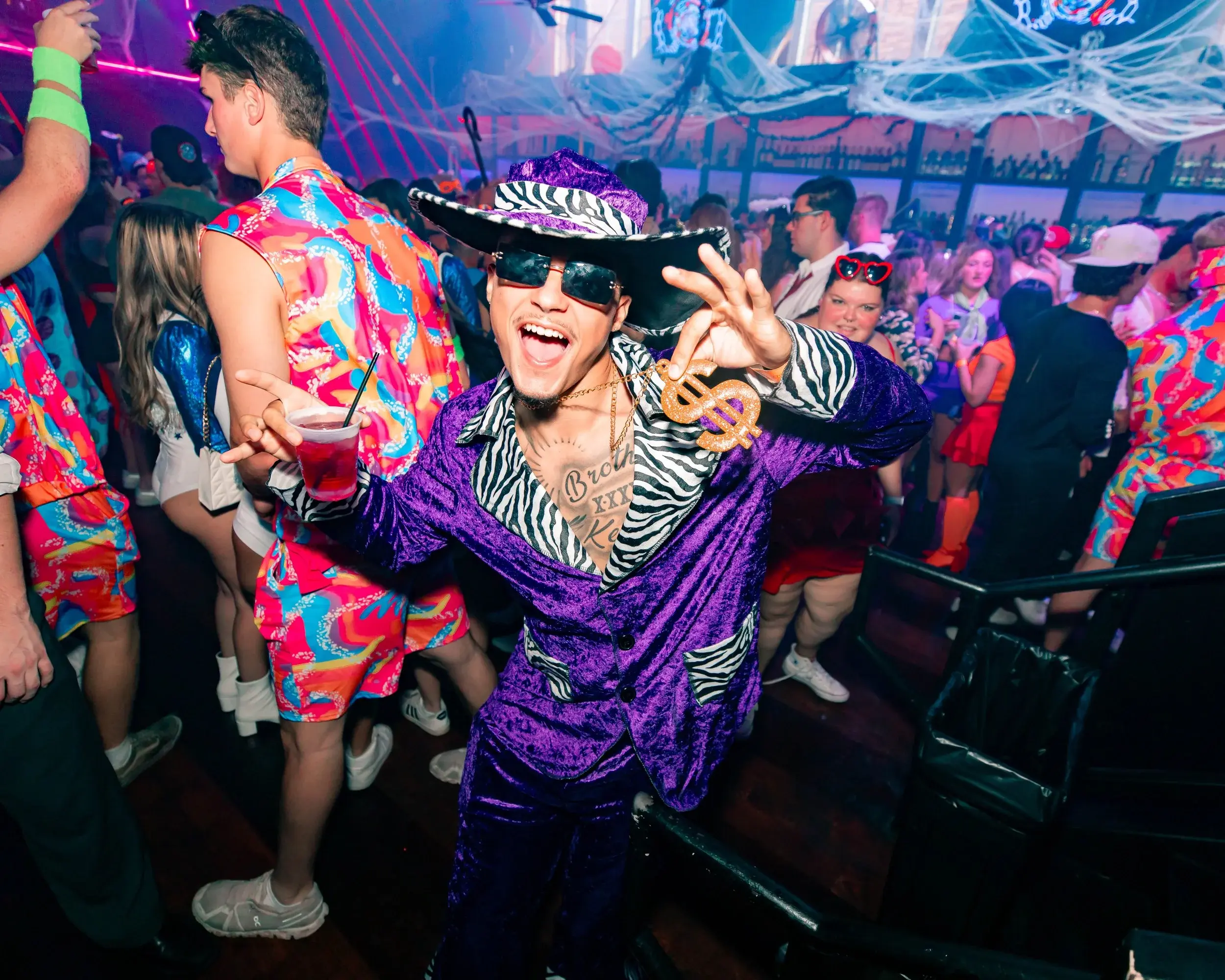 Grinning man in a purple zebra-trimmed costume holding a drink at a colorful costume party