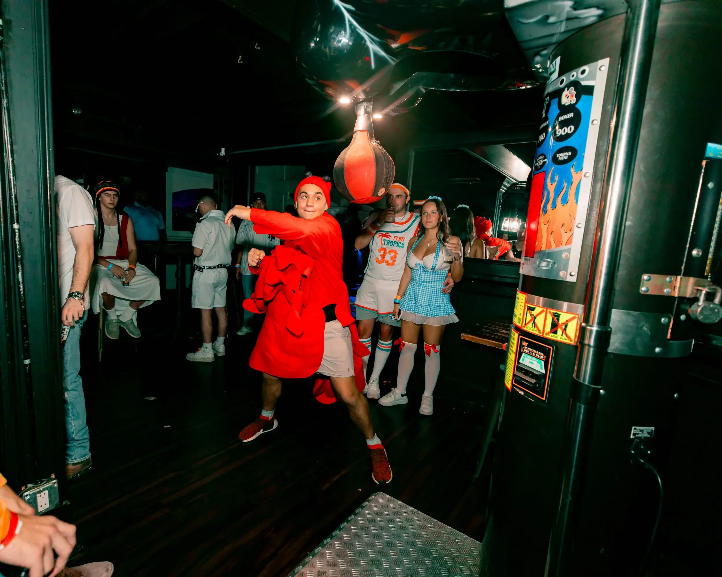 Costumed partygoer in red swinging at an arcade punching bag inside a dark bar