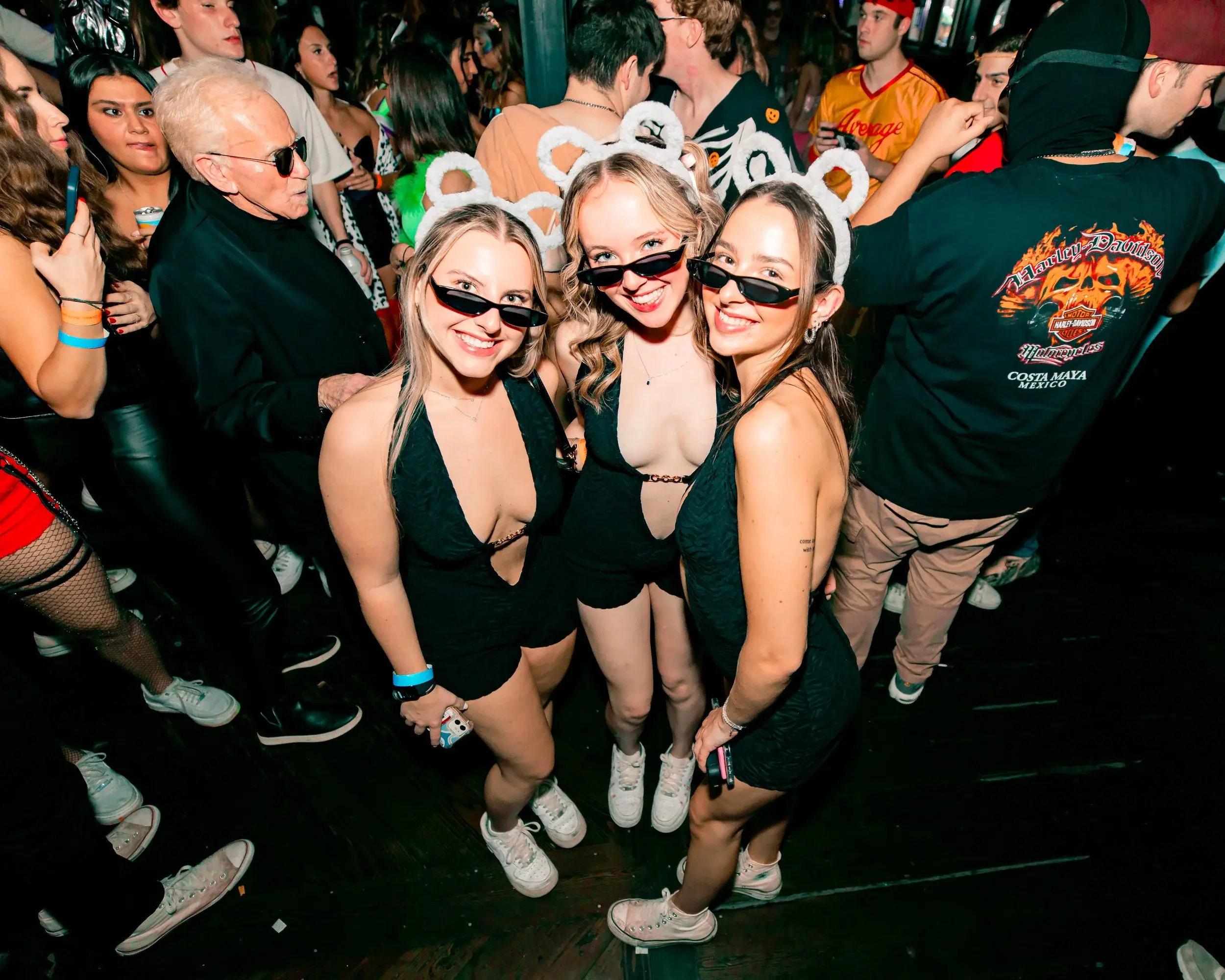 Three friends in black outfits and animal-ear headbands posing on a busy nightlife floor