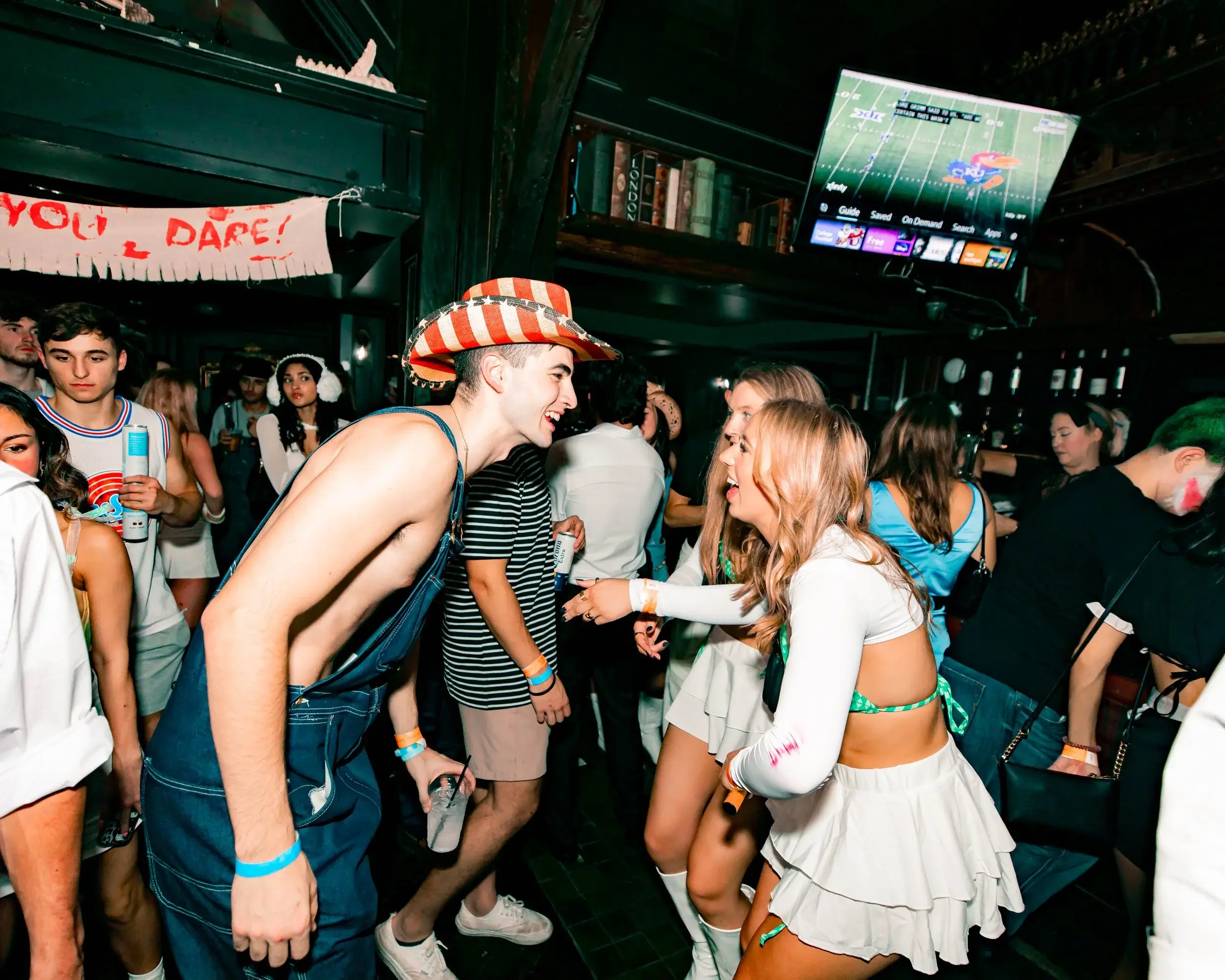 Man in overalls and a straw hat laughing with dancers under a wall-mounted TV