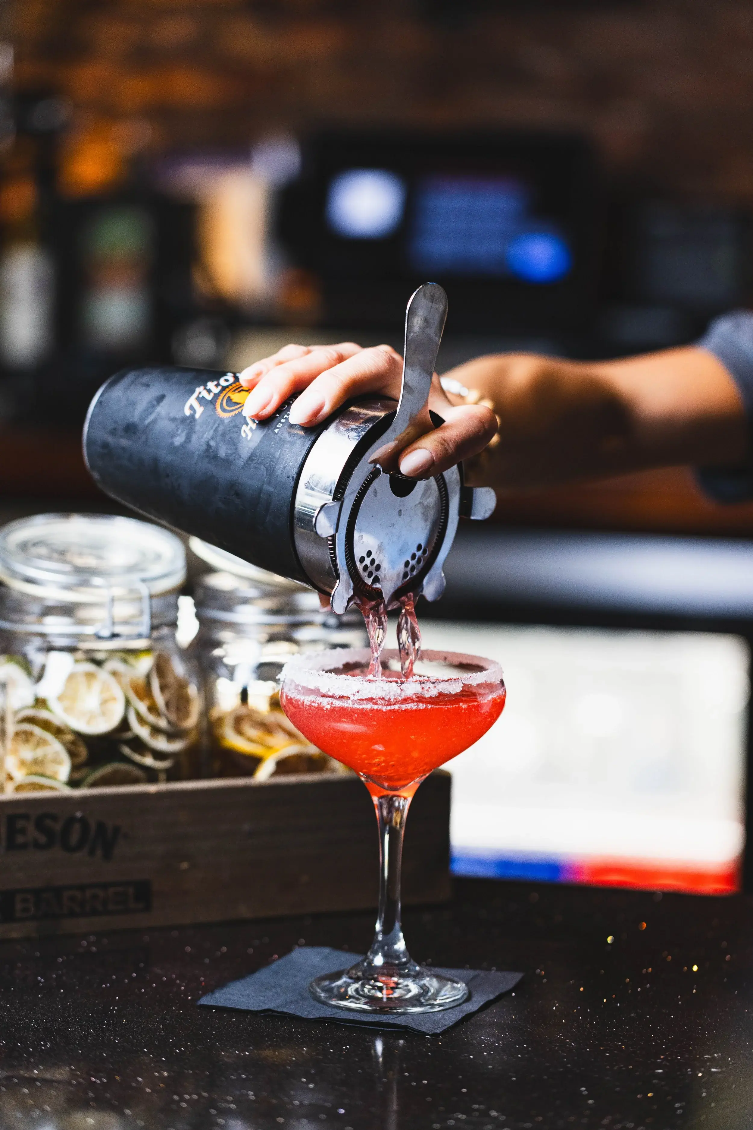 Bartender straining a bright red cocktail into a stemmed glass at the bar