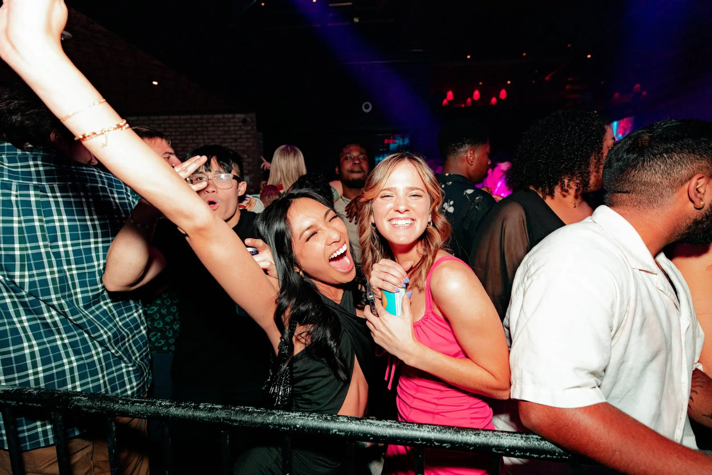 Two friends laughing behind a rail as colored lights sweep the club behind them