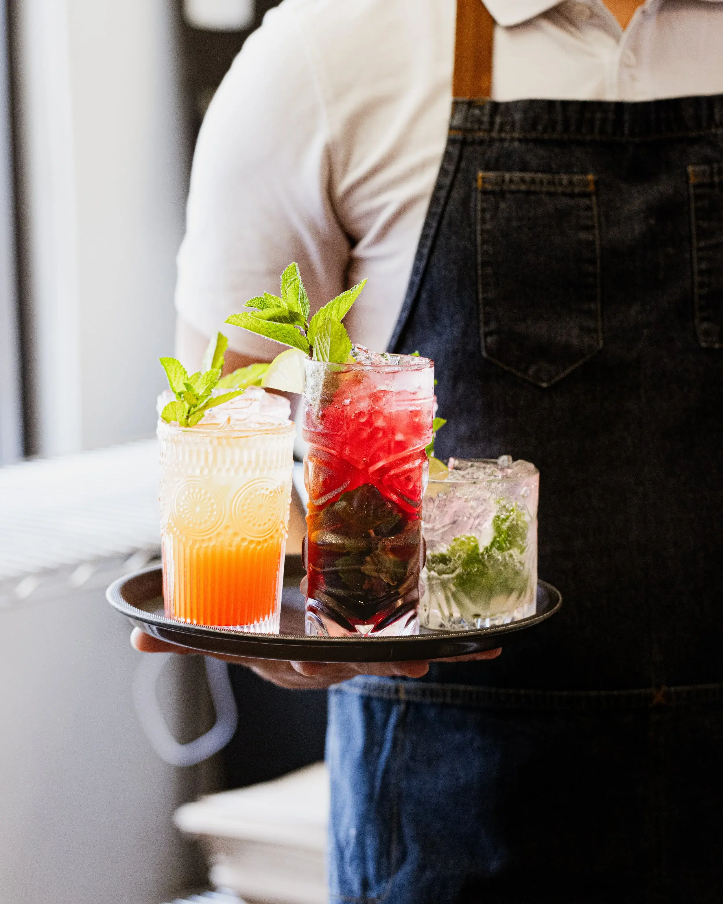 Server in a denim apron carrying a tray of three mint-garnished cocktails
