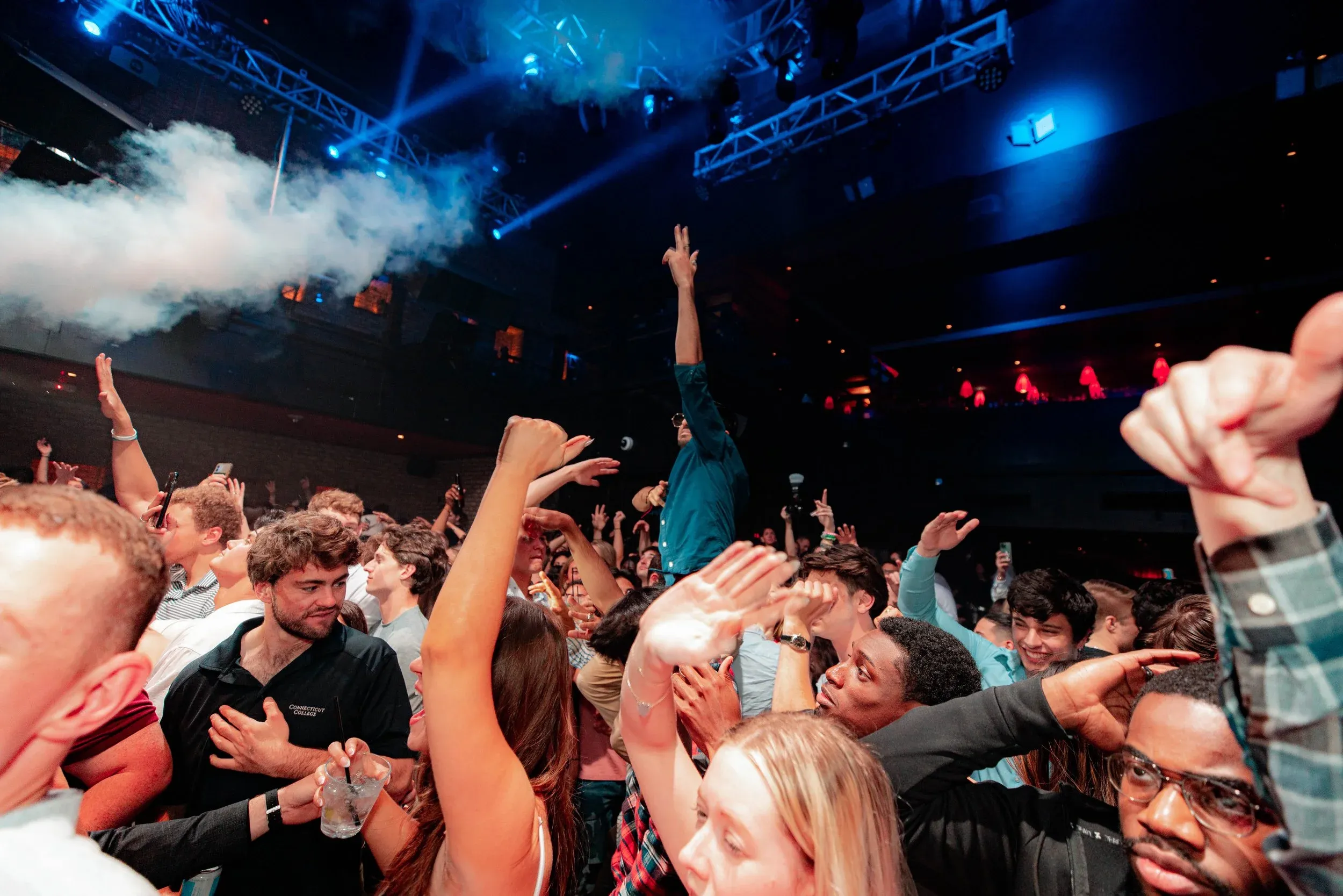 Smoke and blue beams over a packed nightlife crowd with hands raised high