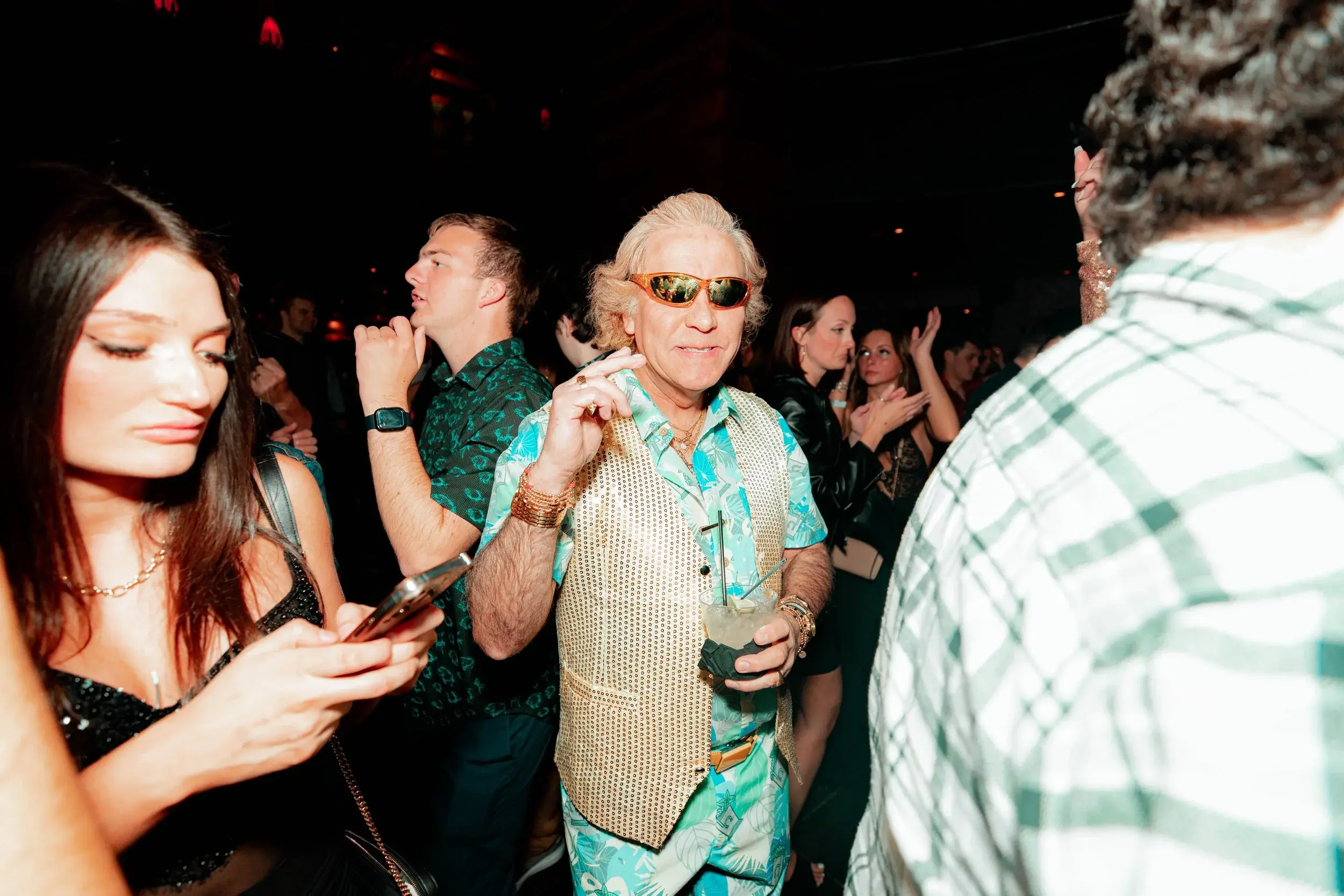 Man in a gold vest and sunglasses holding a drink among nightlife guests