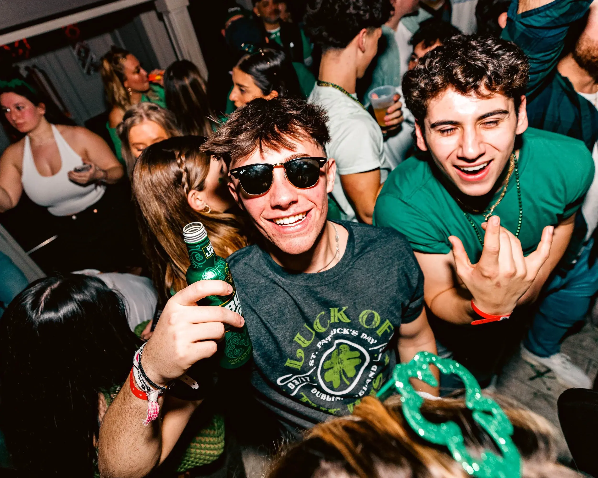 Partygoer in sunglasses grinning with a green bottle beside his friends