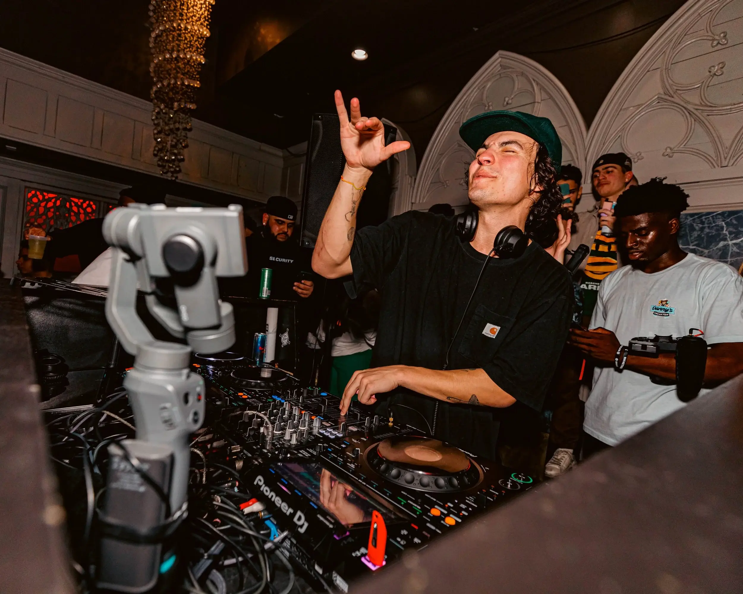 DJ in a green cap raising a hand behind the decks in an ornate white room