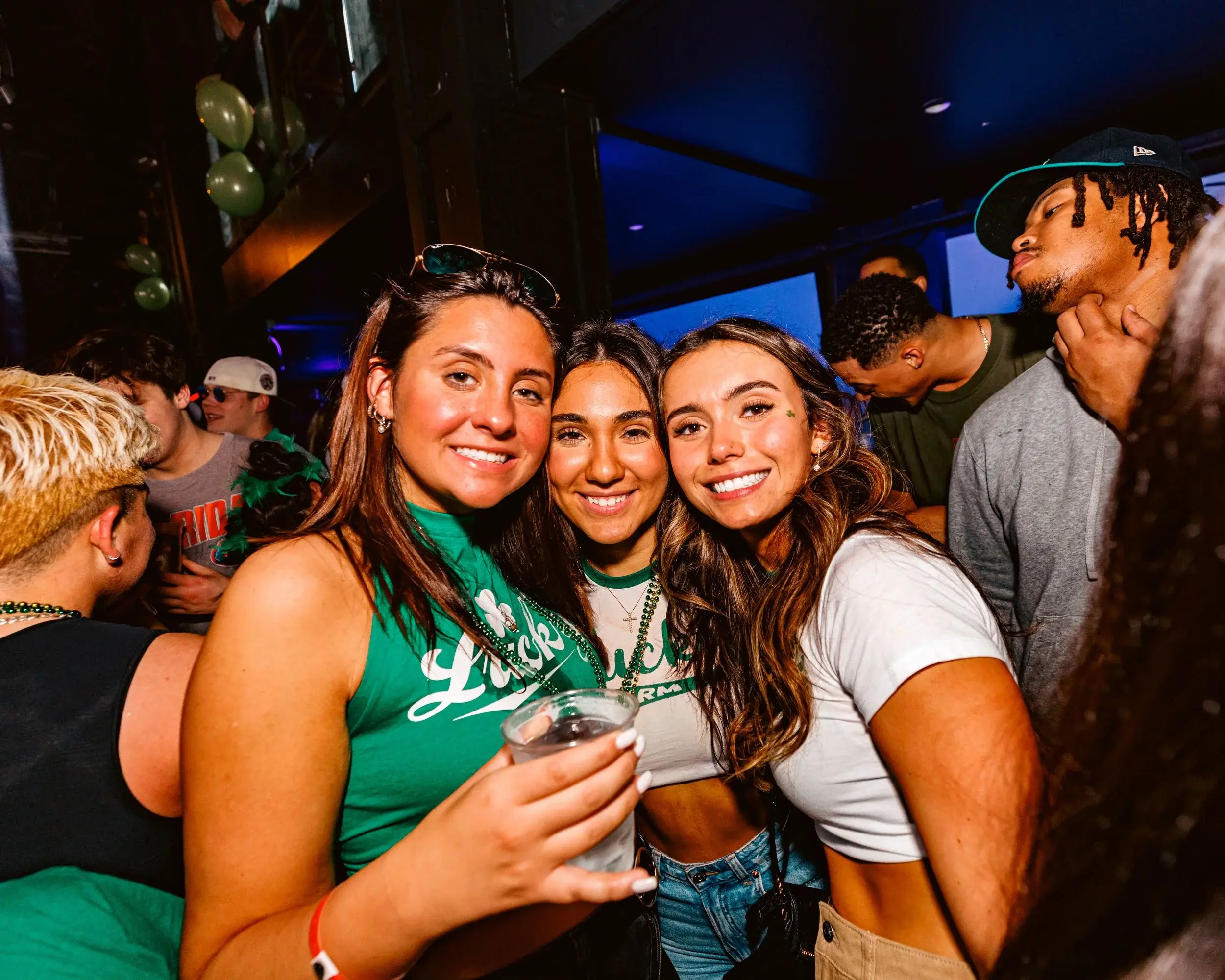 Three friends sharing a drink and smiling beneath green balloons at a bar
