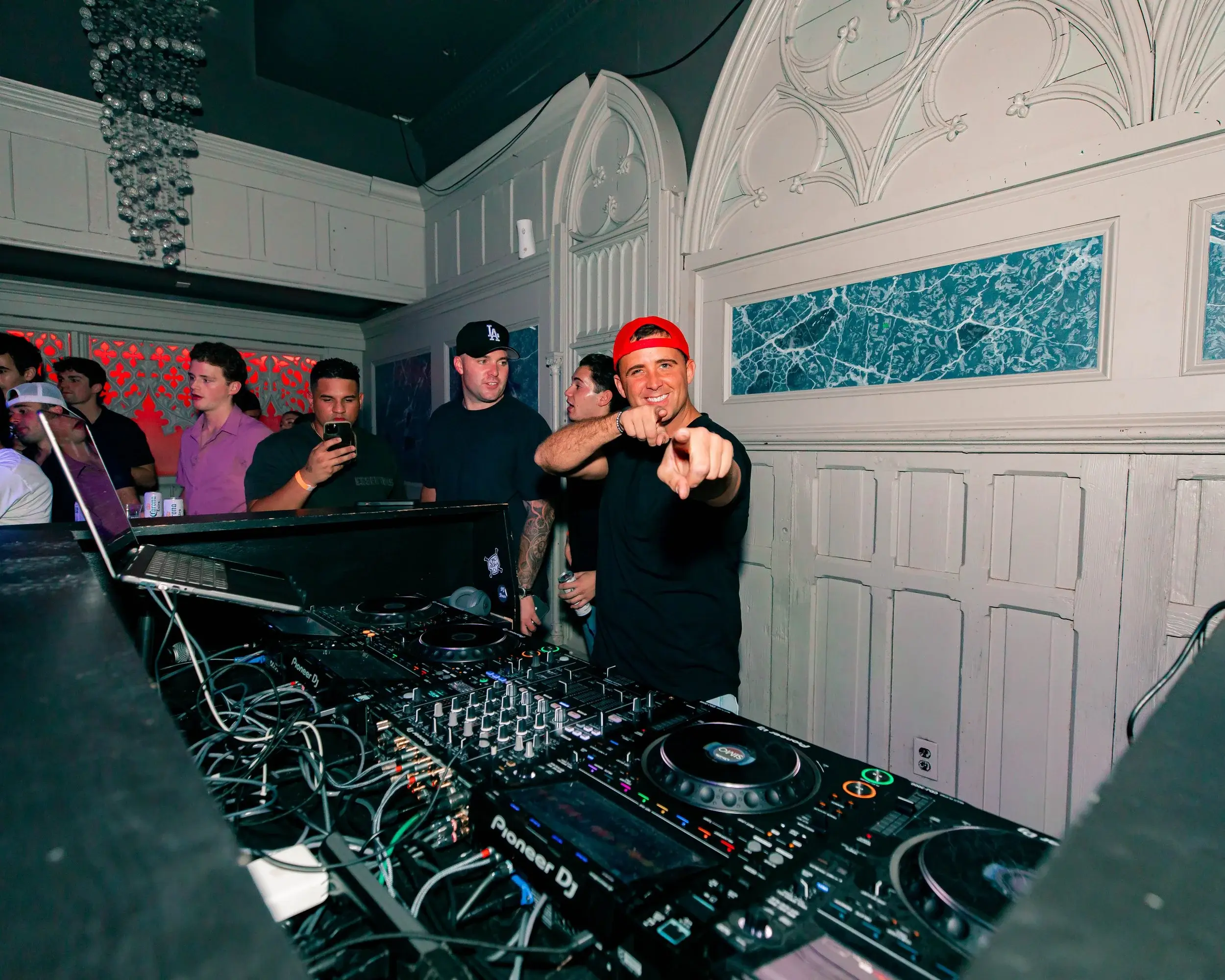 Smiling DJ pointing at the camera from behind a console in a white-paneled room