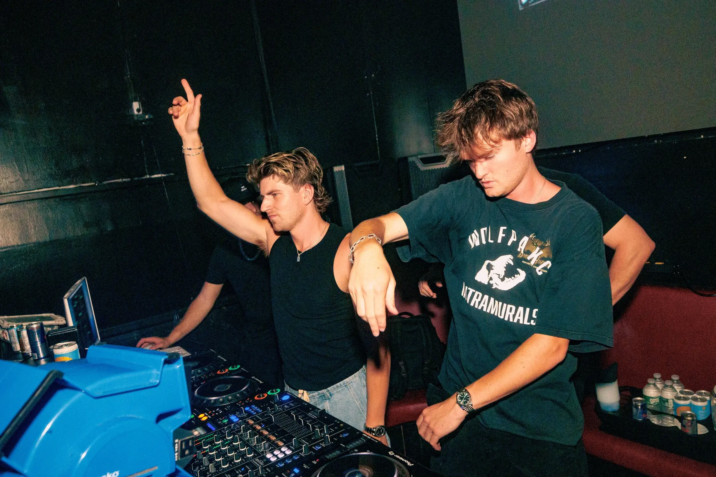 Two DJs working a controller in a dark room, one with an arm raised