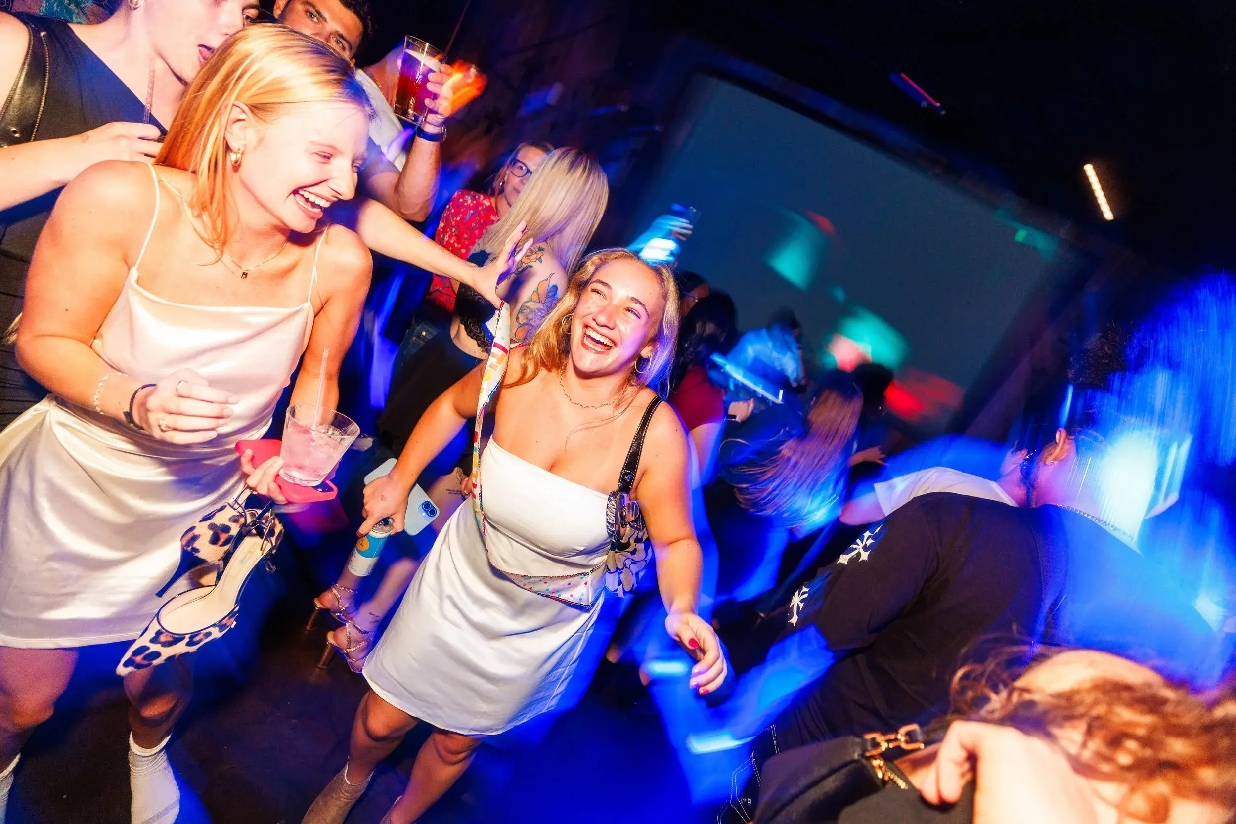 Two friends in white dresses laughing while dancing under deep blue club lights