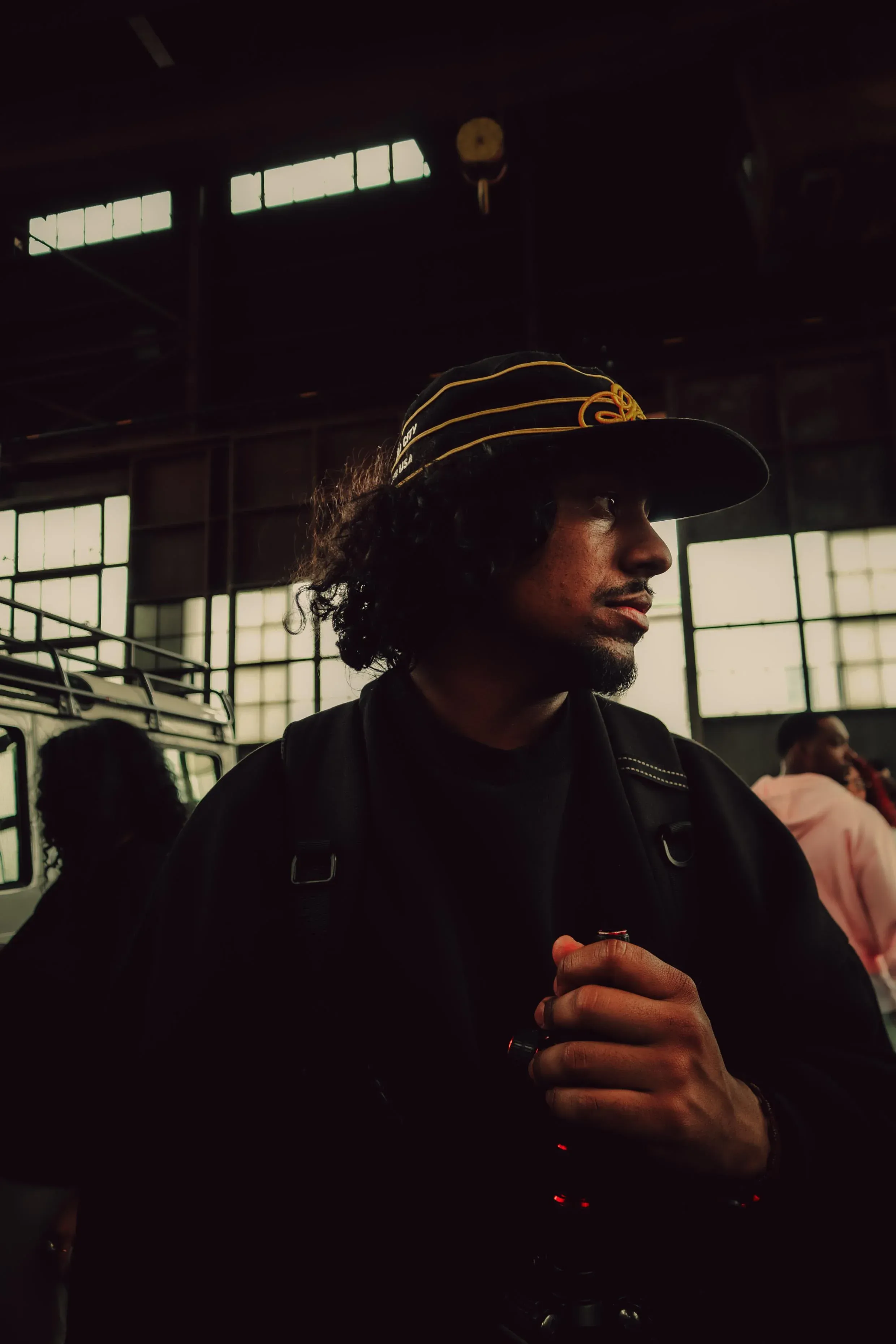 Photographer Mack Minaya in a dark jacket and embroidered cap inside an industrial venue with skylights