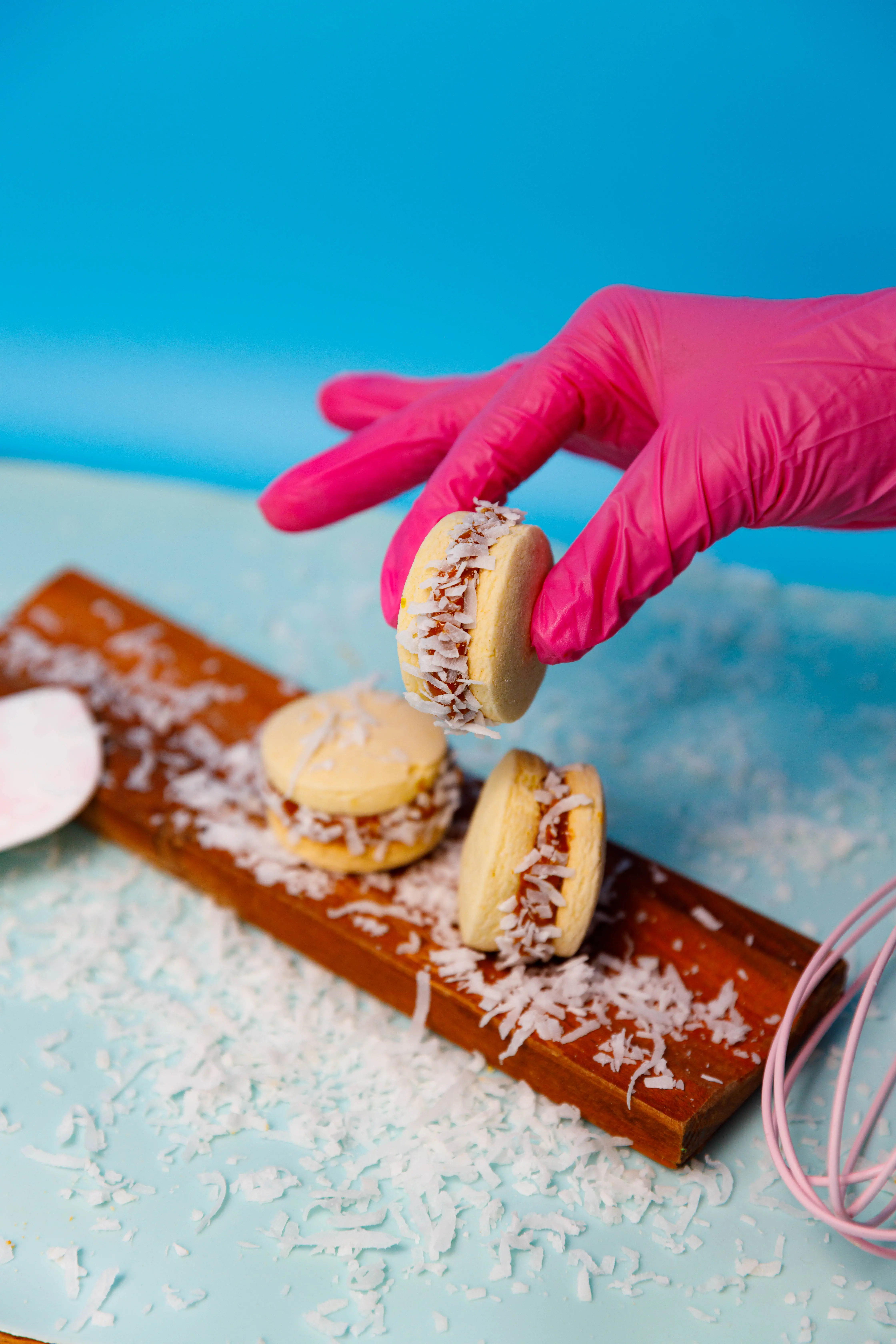 Pink-gloved hand stacking coconut-dusted alfajores on a board against a blue backdrop