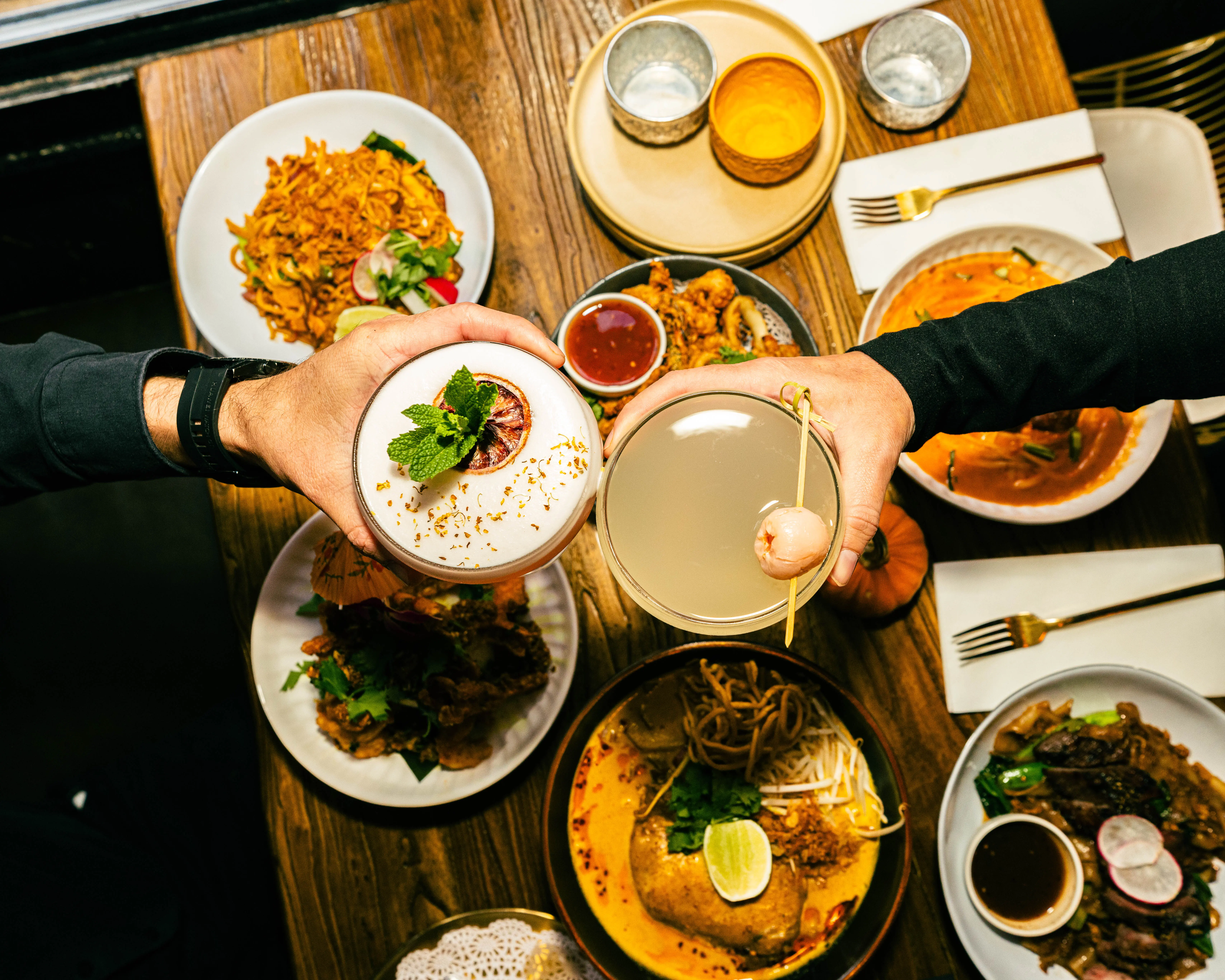 Two guests toasting cocktails above a table of shared dishes