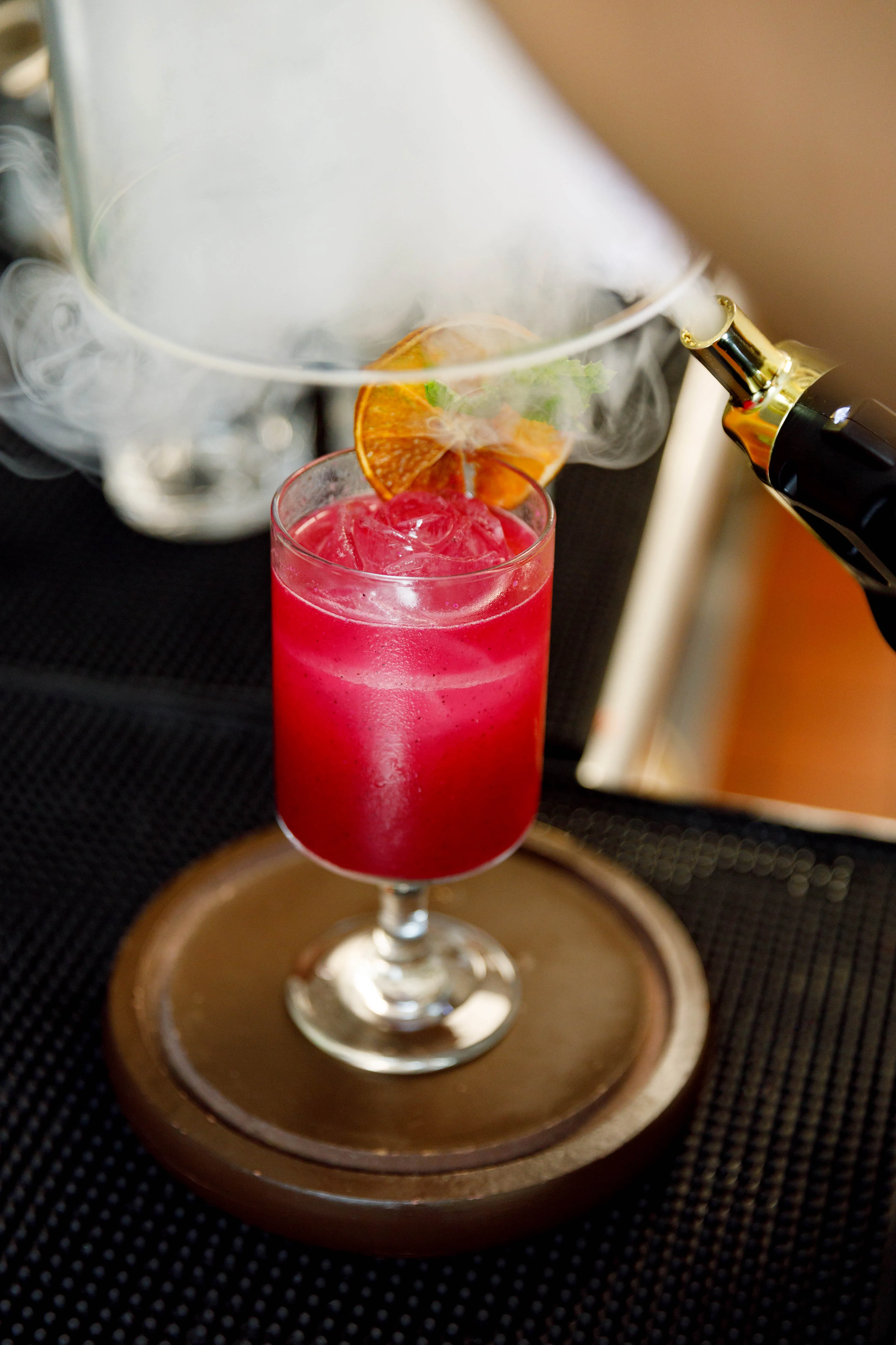 Smoke drifting over a red cocktail with a dried orange wheel at the bar