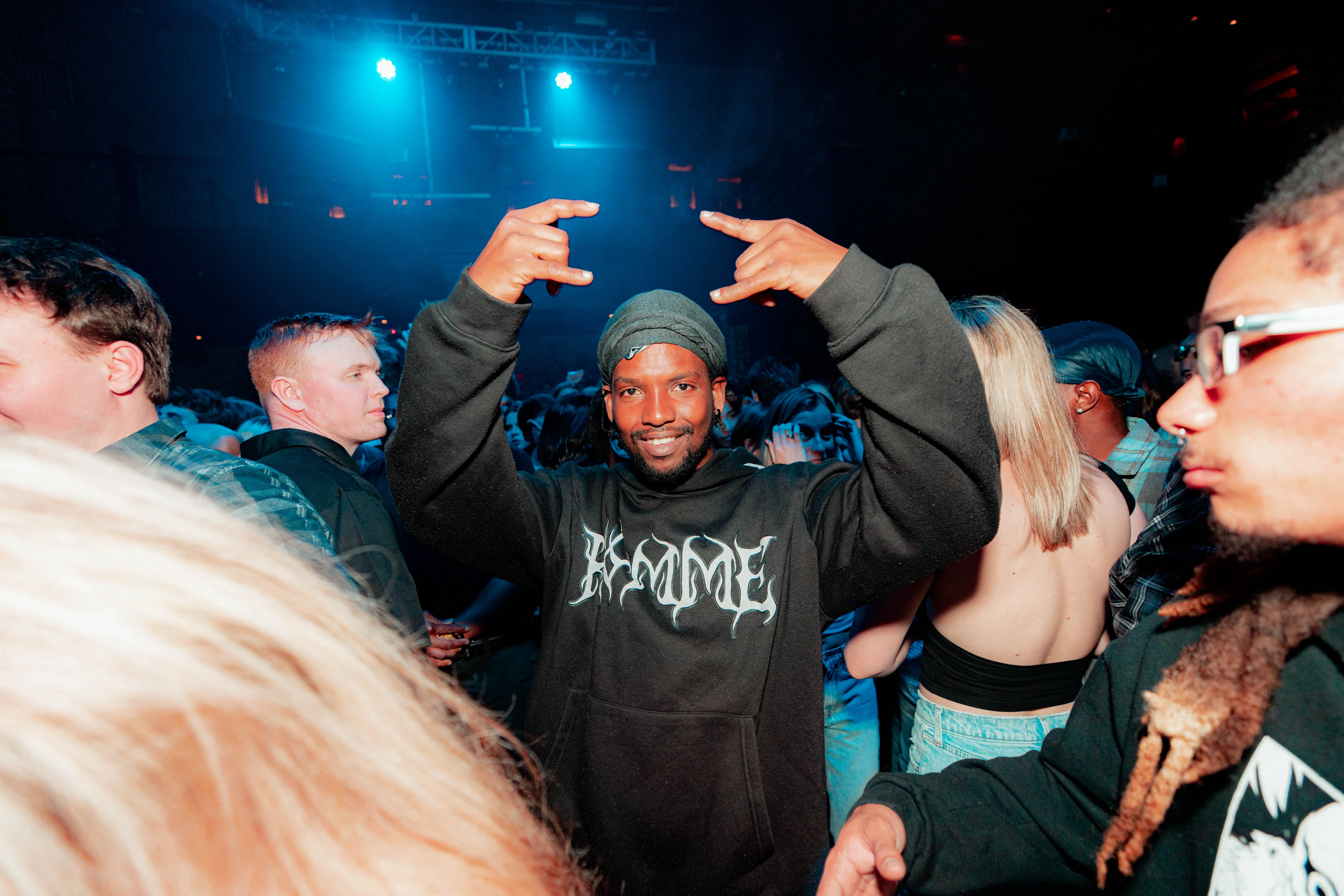Man in a beanie throwing hand signs in a blue-lit concert crowd