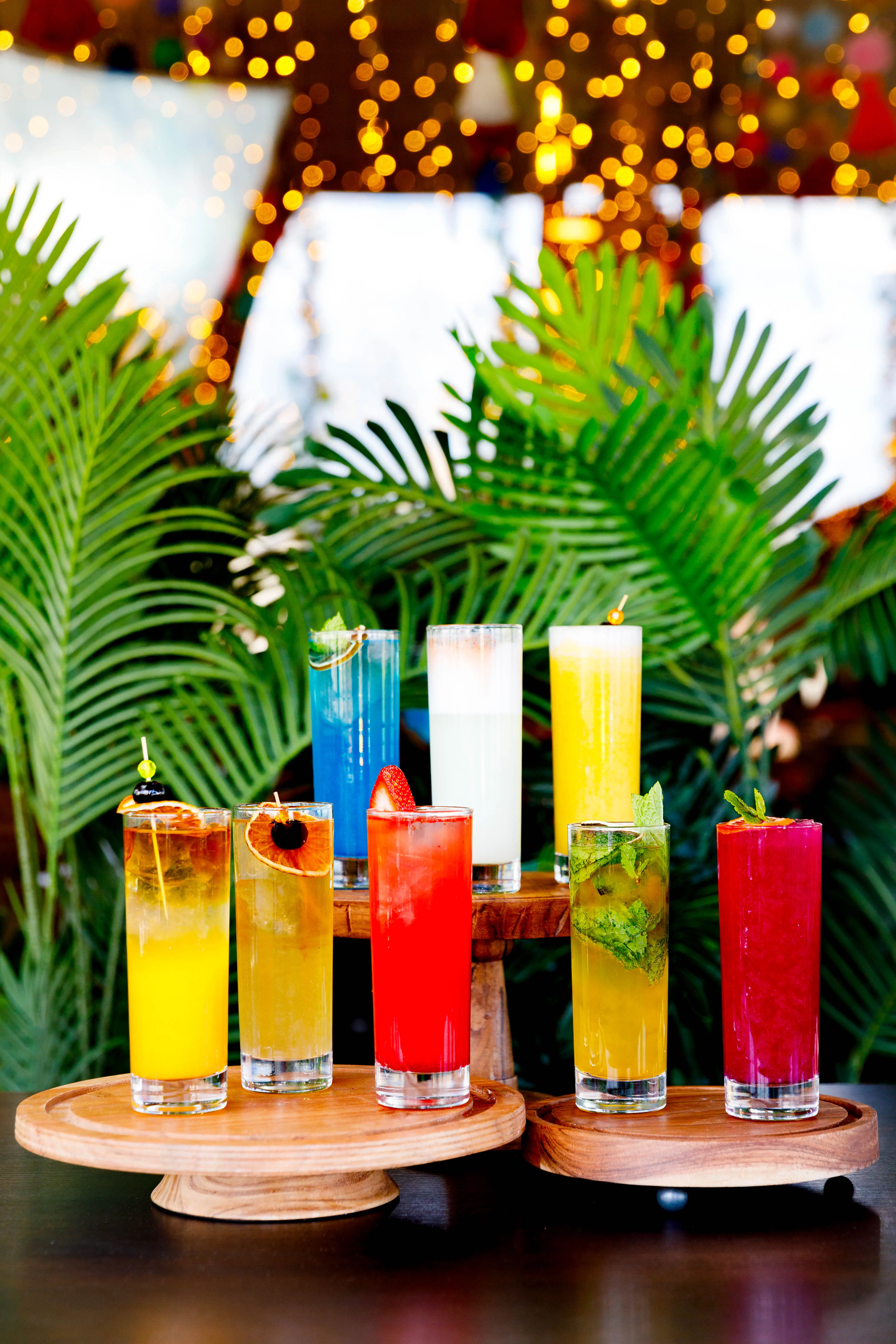 Row of colorful cocktails garnished with fruit and flames against palm leaves