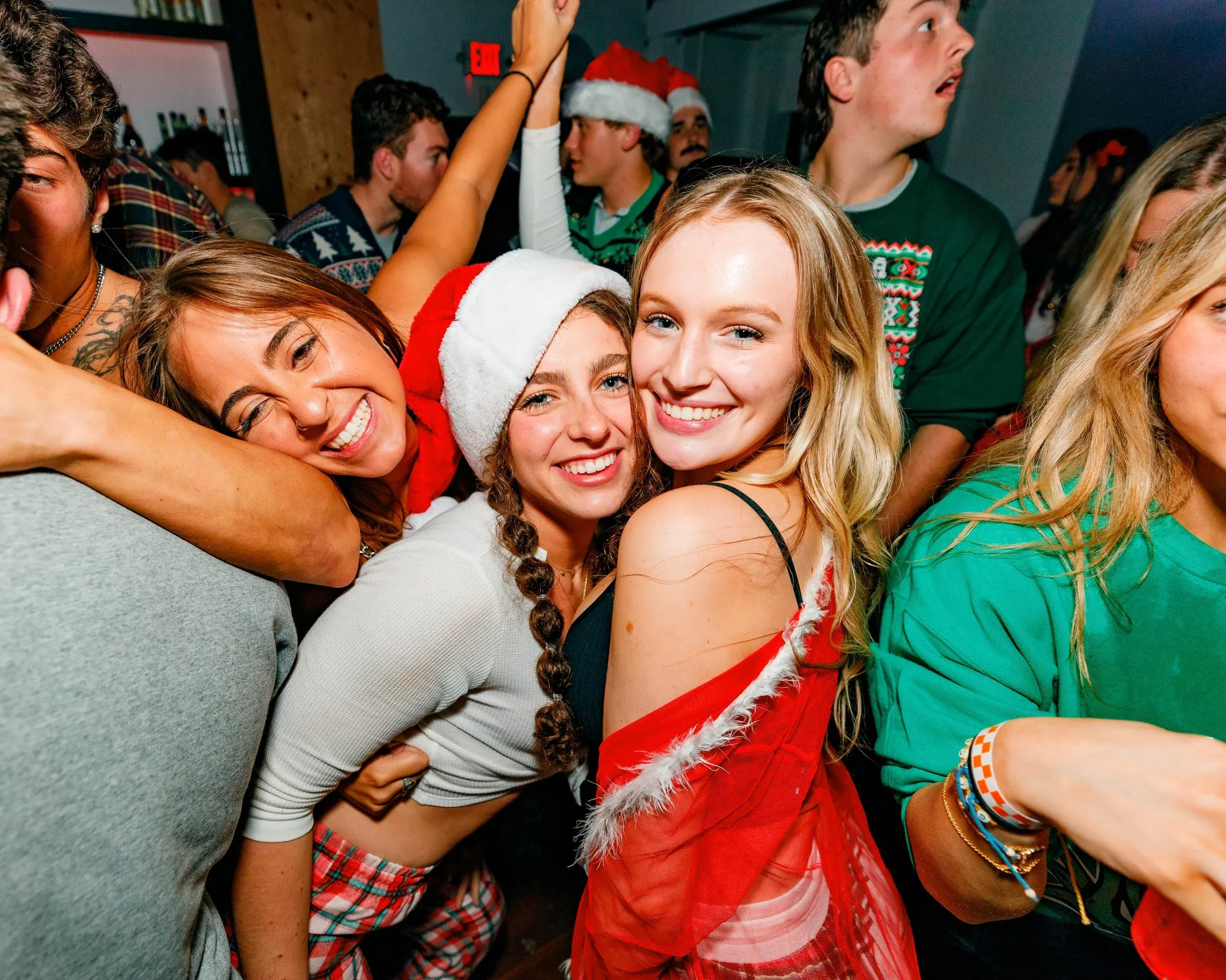 Smiling friends in Santa hats embracing in a crowded room at a holiday celebration