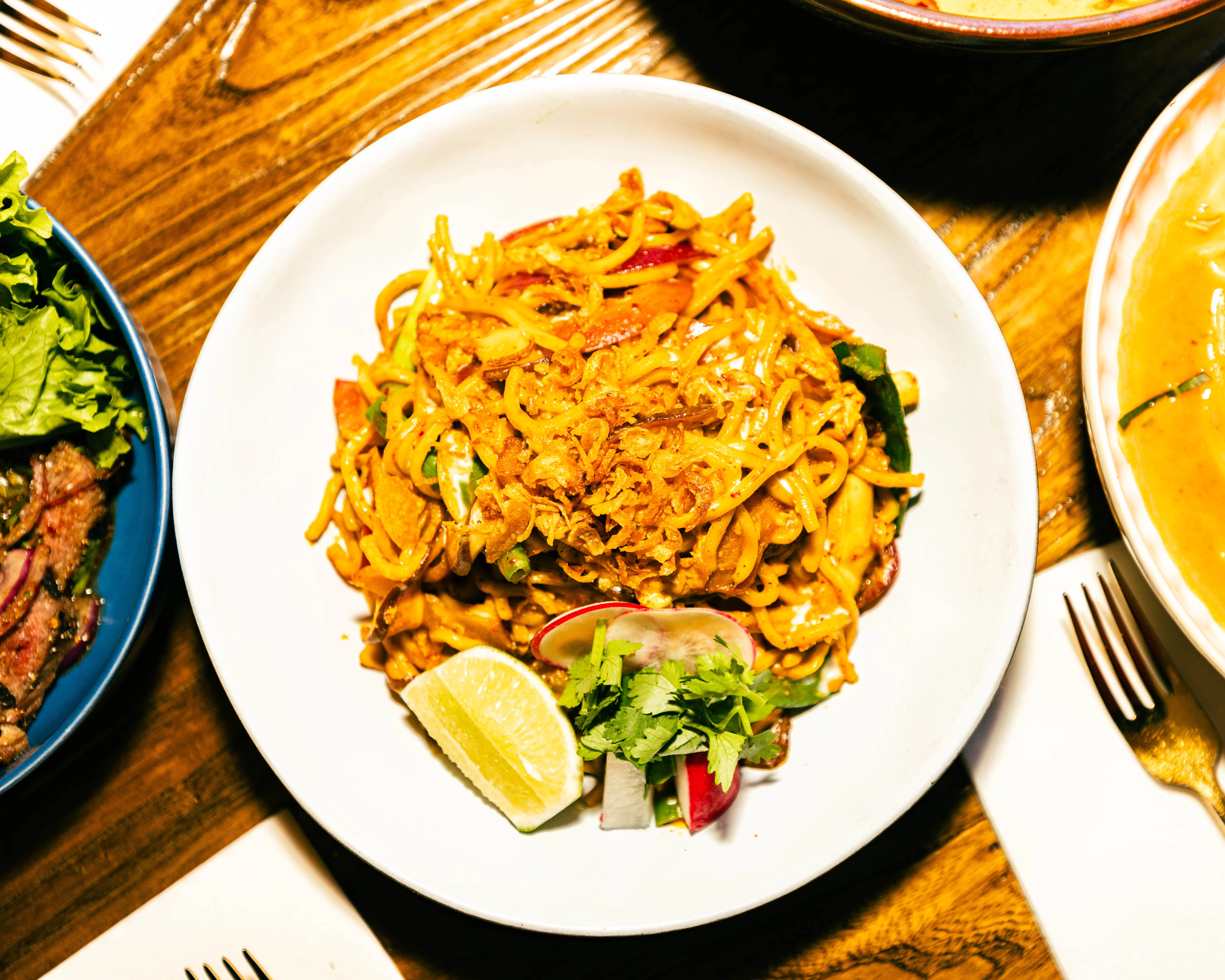 Stir-fried noodles topped with crispy shallots, lime, and microgreens on a set table