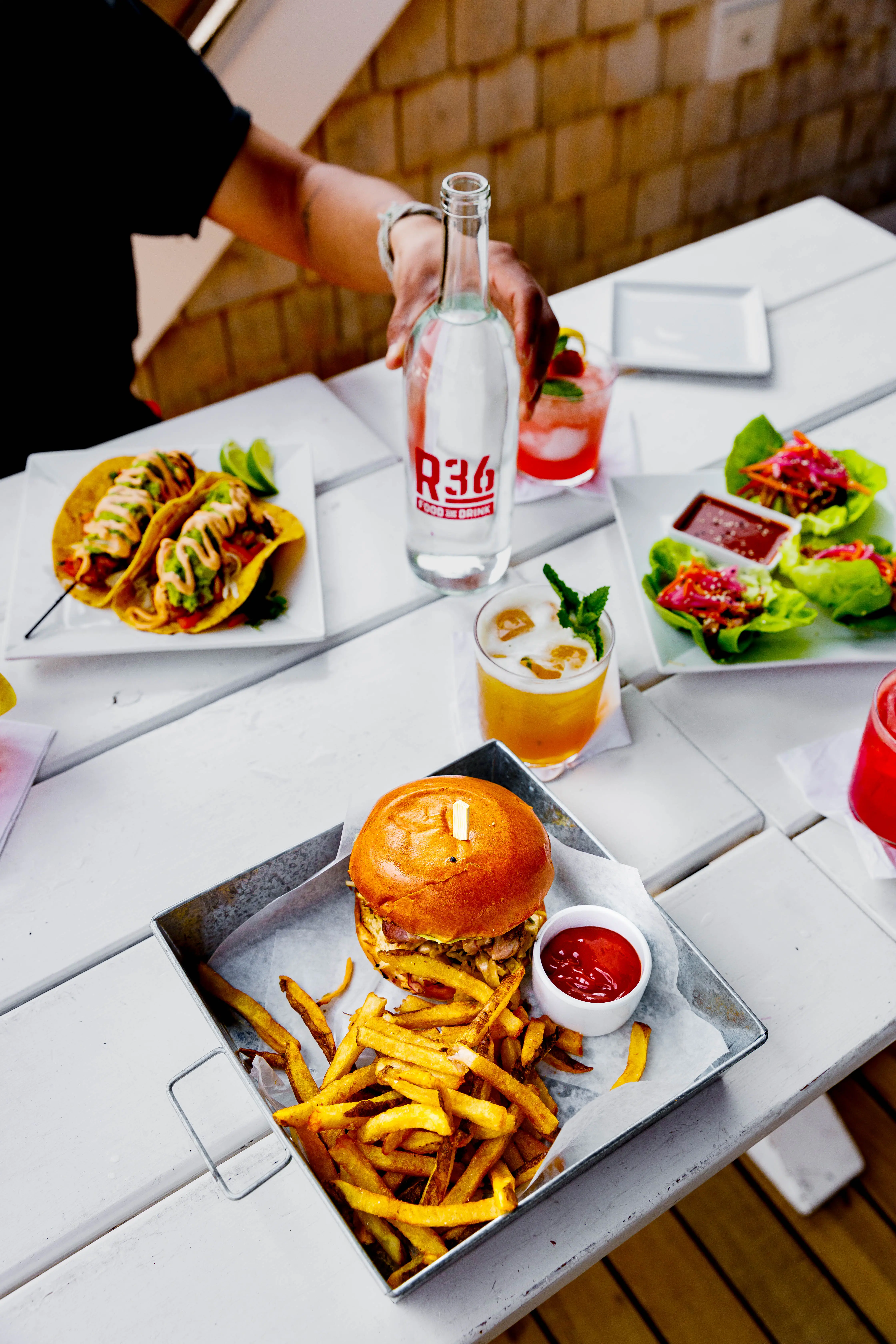 Outdoor table set with a burger, fries, tacos, and cocktails