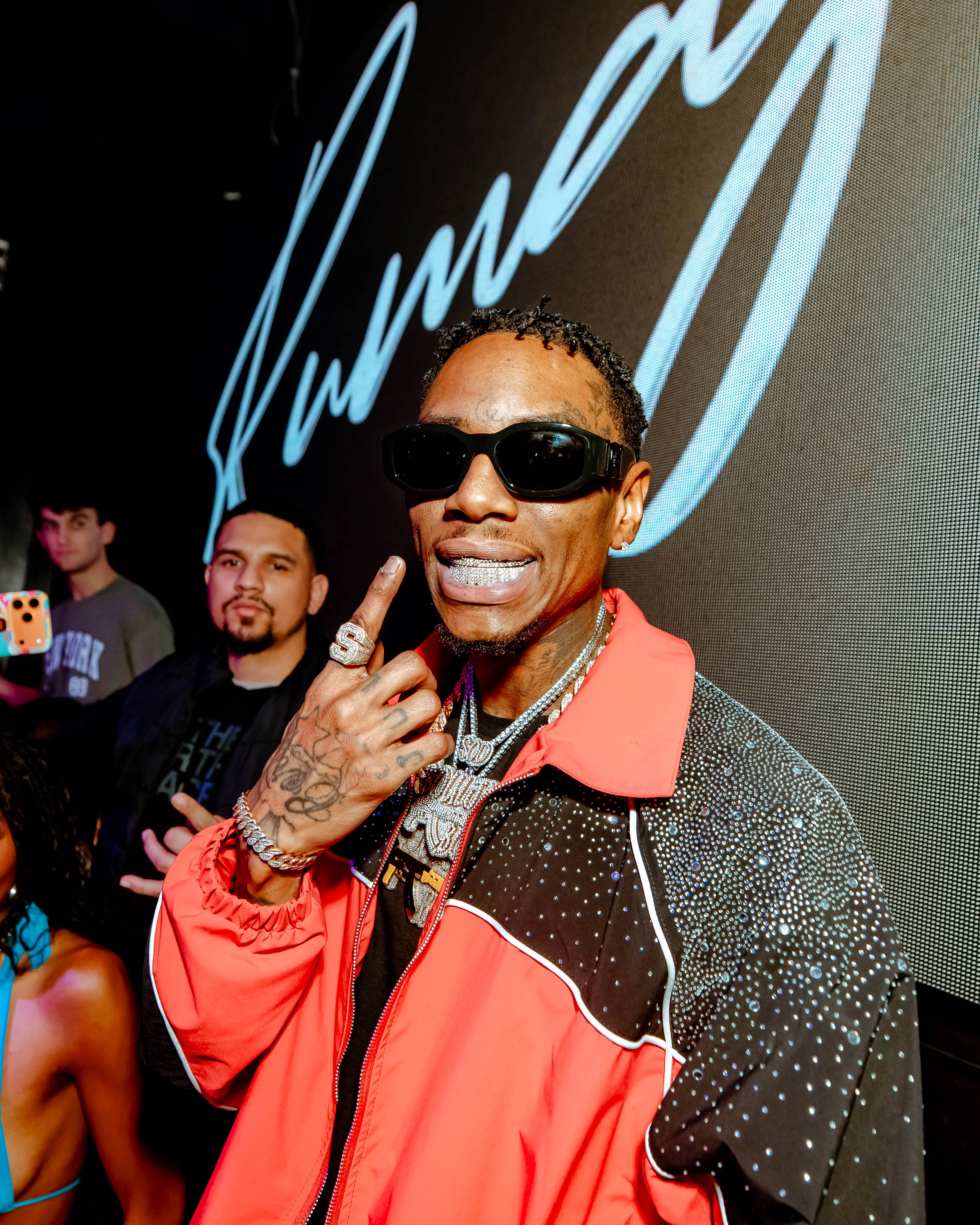 Guest in a rhinestone-studded jacket and sunglasses flashing jewelry at a club appearance