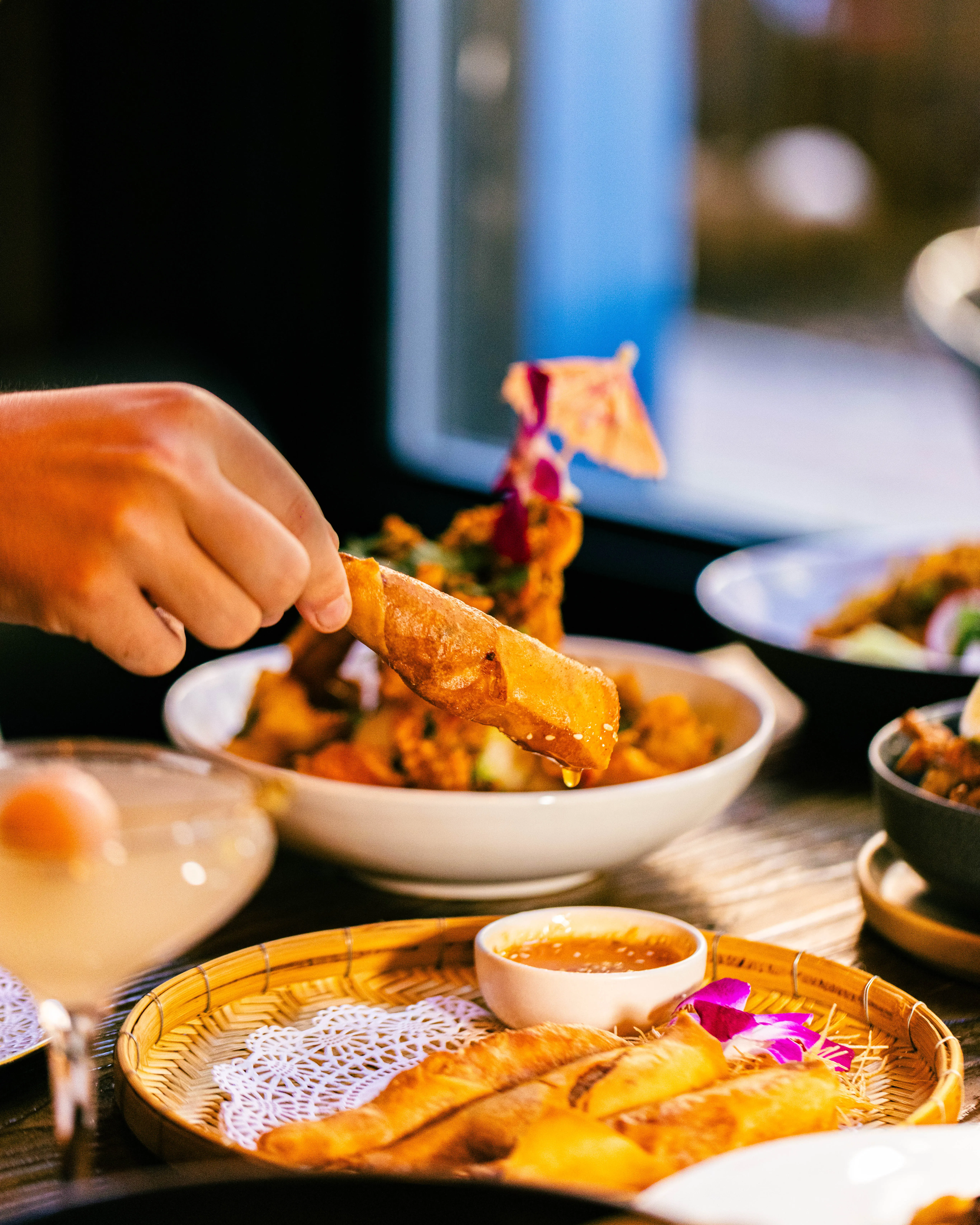 Hand lifting a crispy spring roll over plated dishes at a restaurant table