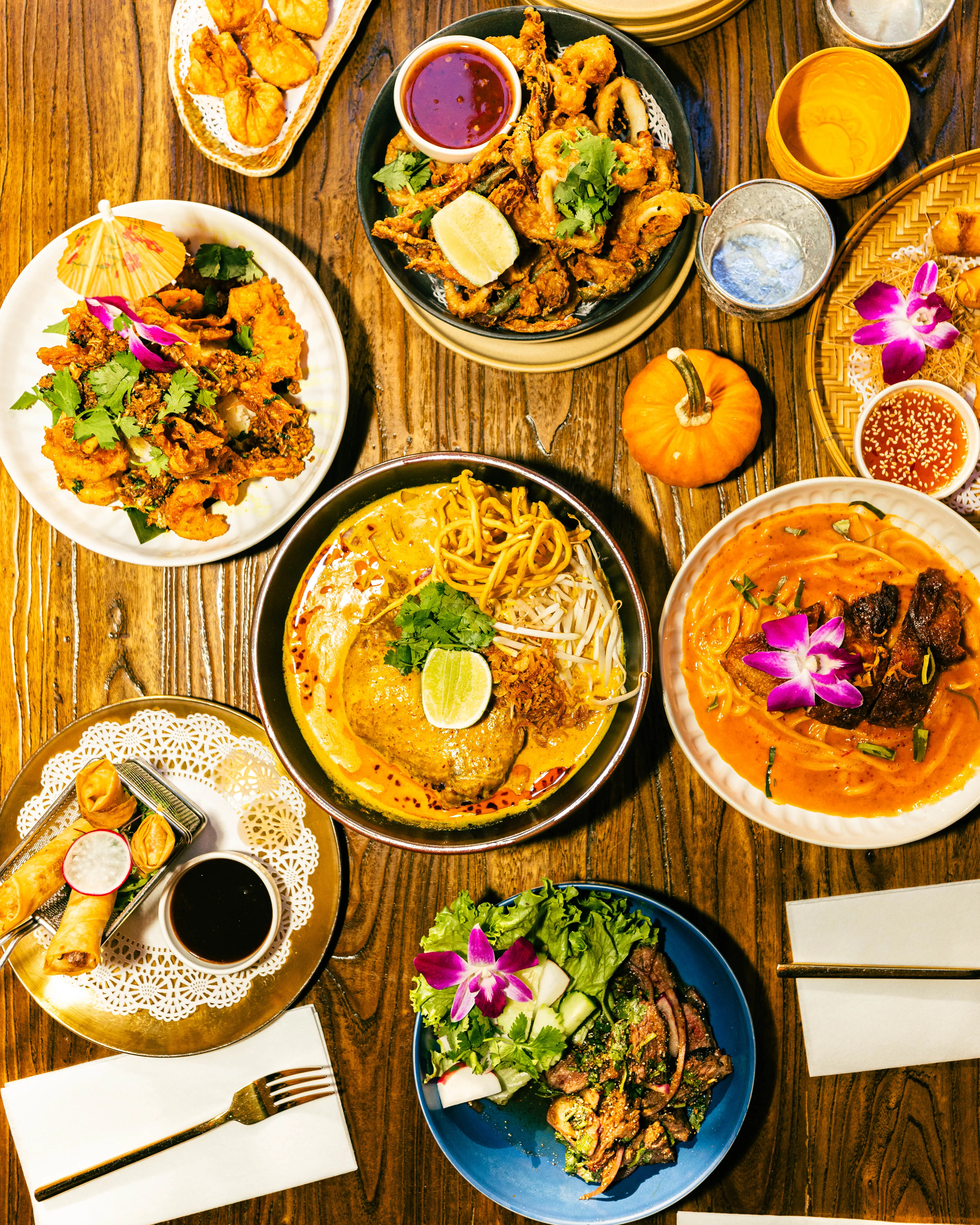 Overhead restaurant table crowded with noodle bowls, curries, and fried appetizers